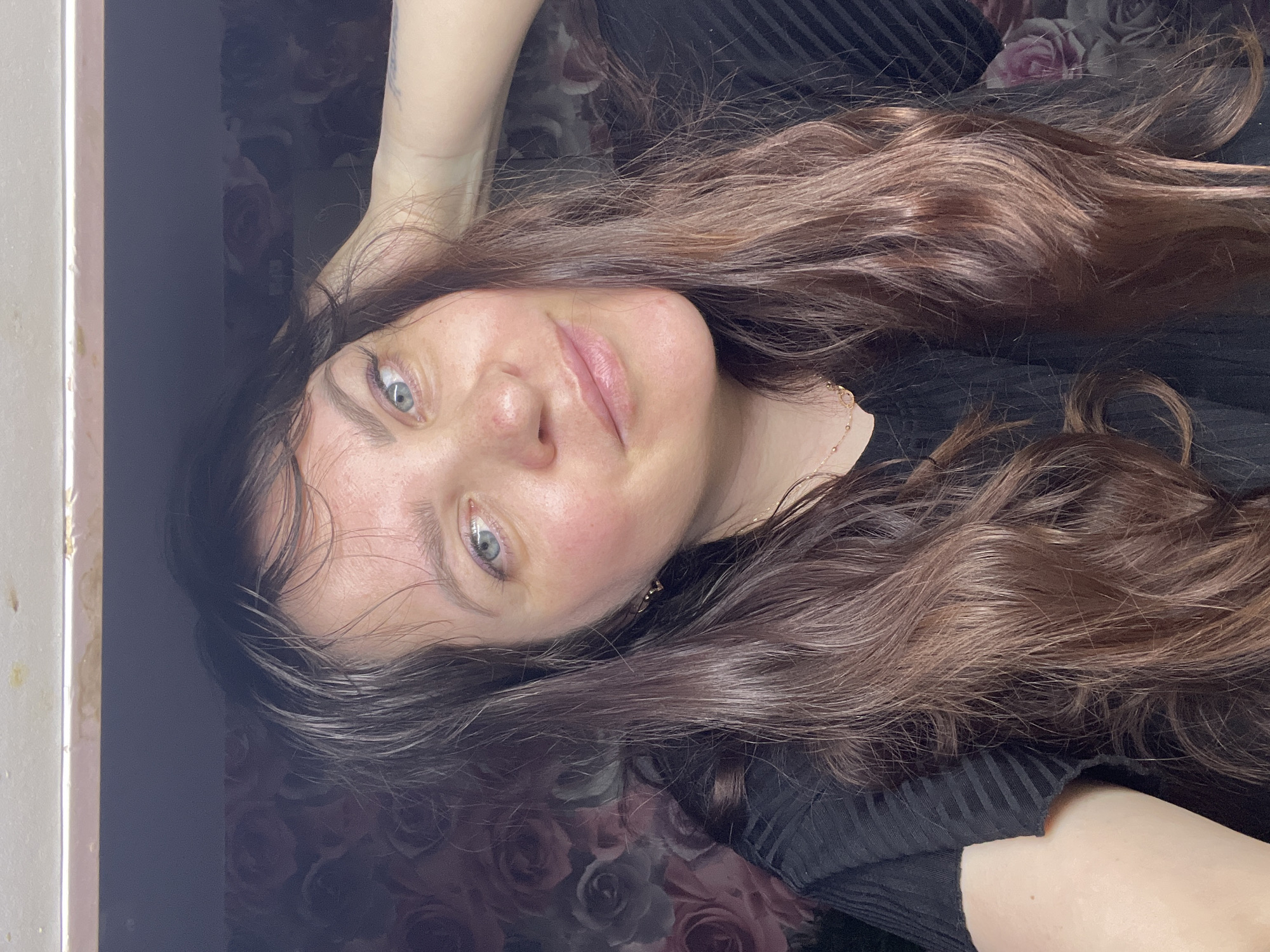 The image shows a close-up, selfie-style shot of a woman with long, wavy brown hair and light blue eyes. She is looking towards the camera with a slight smile. Her right hand is raised to the back of her head. She is wearing a black ribbed top and a delicate gold necklace. The background appears to be a wall with a floral wallpaper featuring roses in shades of pink and gray. A portion of a white shelf or fixture is visible at the top of the image, with some small yellow stains on it. The lighting suggests an indoor setting, possibly a room in a residential building. The image does not contain any specific clues about its location in Noviy, Russia.