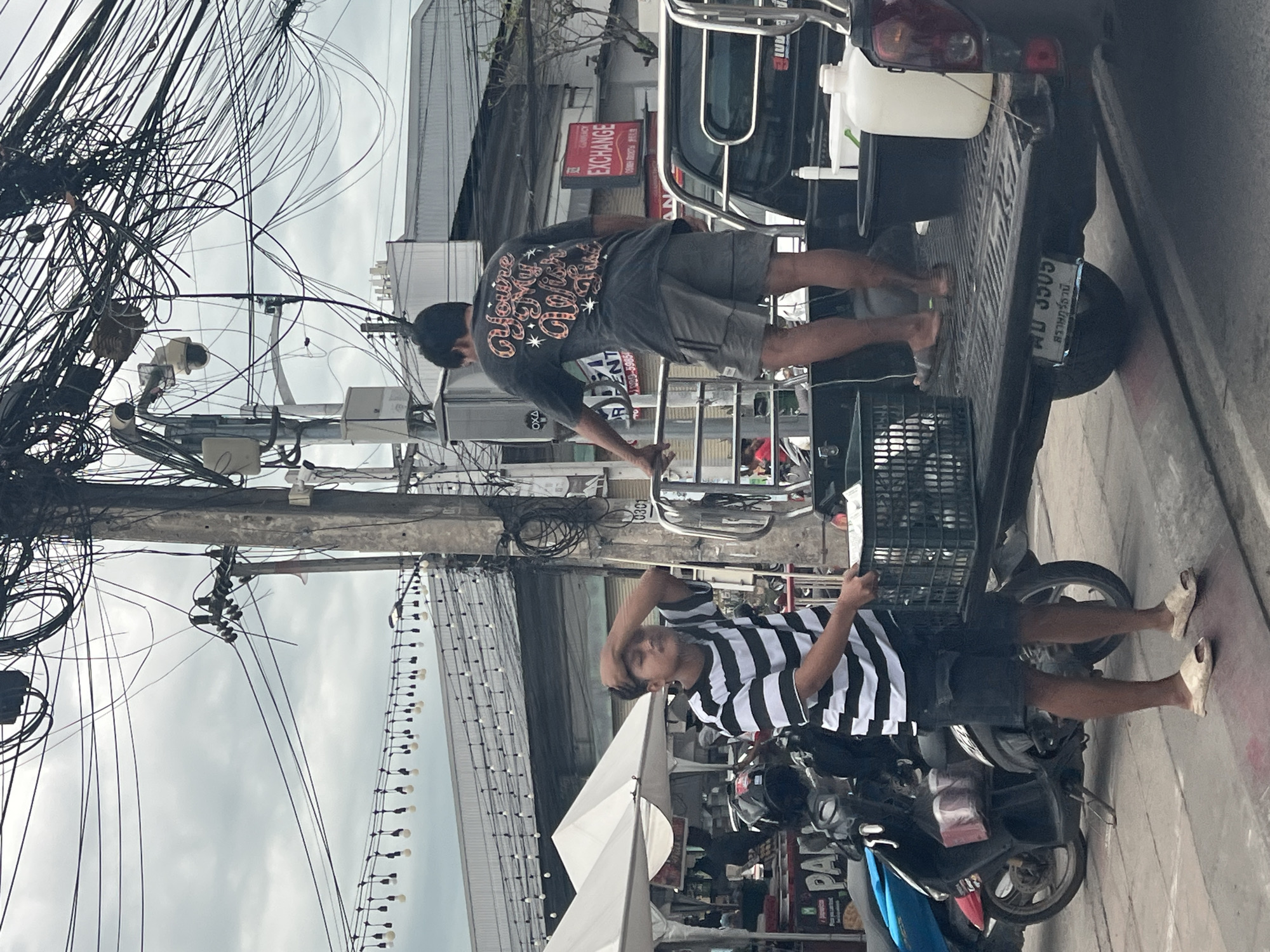 A young man wearing a black and white striped t-shirt and dark shorts, with his hand on his forehead, stands on a sidewalk in front of a parked motorcycle and a pickup truck in Ban Bang Rak, Thailand. He is holding a black plastic crate. Behind him, a man in a dark t-shirt and gray shorts stands on the bed of the pickup truck, leaning on the railing. The truck bed contains a large white plastic container and a black tub. The scene is characterized by a dense tangle of overhead electrical wires and streetlights. In the background, buildings with corrugated metal roofs and a sign for "EXCHANGE" are visible. The sky is overcast.