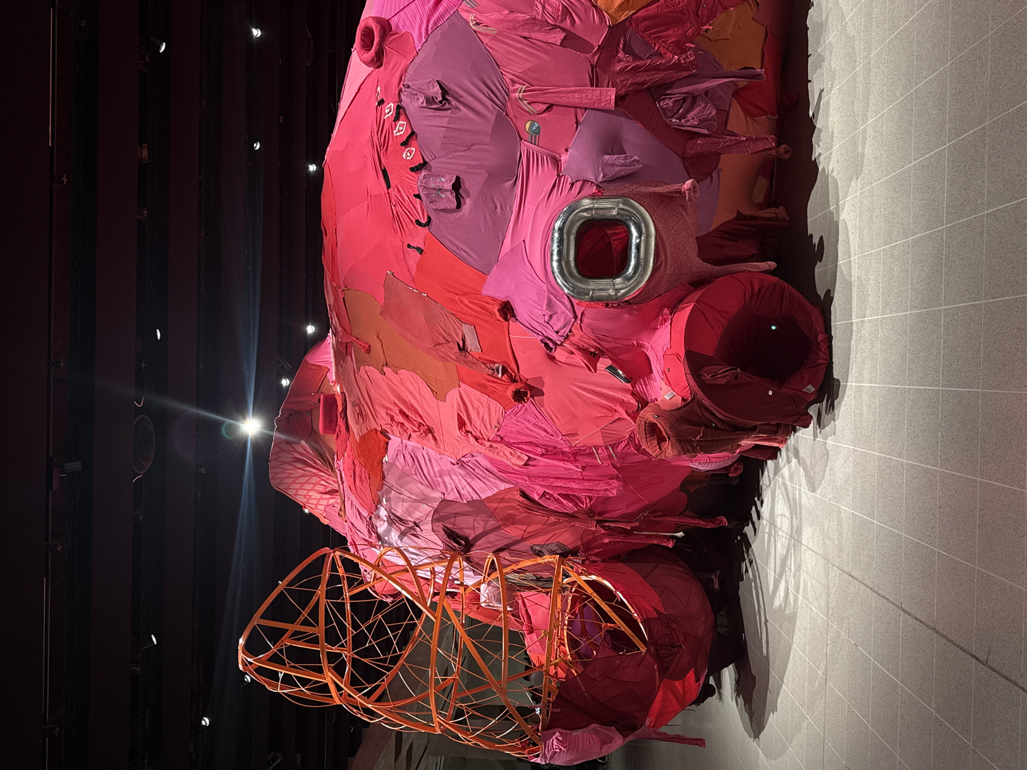 The photograph depicts a large, abstract sculpture primarily composed of red and pink fabric scraps, possibly clothing remnants. A distinctive silver metal ring is embedded in the fabric on the sculpture's upper right side. Underneath, the sculpture includes a cage-like structure of orange rods. The sculpture casts a shadow on a gray tiled floor. The background is dark, with visible lighting fixtures, suggesting an indoor exhibition space. The sculpture is located at the Hayward Gallery in the city of London, United Kingdom.