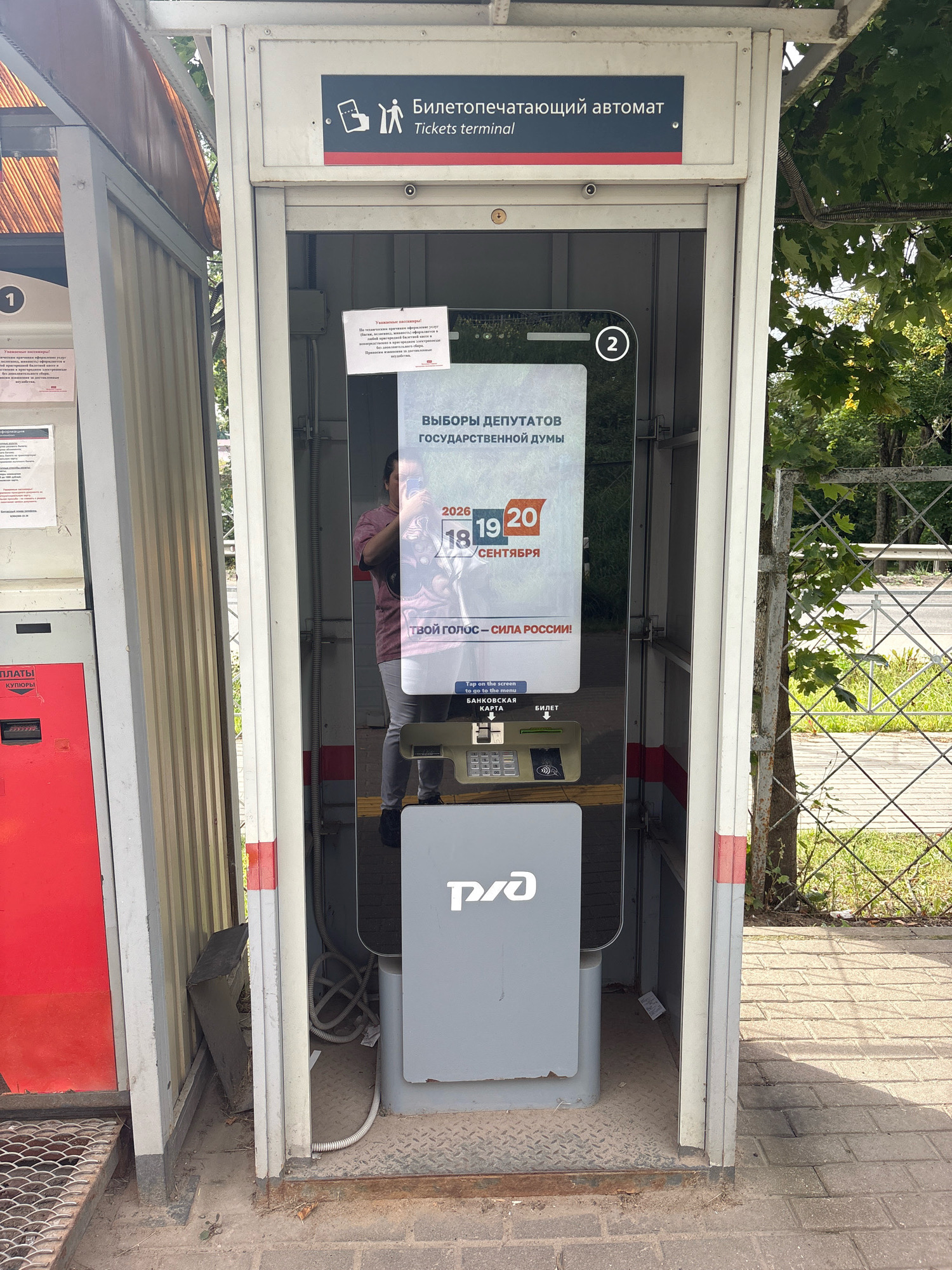 An outdoor ticket vending machine is situated next to a sidewalk and a fenced area with trees. The machine is housed in a booth with a transparent front, allowing visibility of its interior. The booth has a sign at the top that reads "Билетопечатающий автомат Tickets terminal." Inside the booth, a screen displays information related to elections, specifically mentioning "ВЫБОРЫ ДЕПУТАТОВ ГОСУДАРСТВЕННОЙ ДУМЫ" and dates "18-20 СЕНТЯБРЯ." Below the screen, a card reader and a bill acceptor are visible. A person's reflection can be seen in the screen, holding a phone. To the left of the booth is a red machine with the word "ОПЛАТЫ" visible. The ground around the machine is paved with bricks and metal grating. The overall environment appears to be an urban or semi-urban setting.