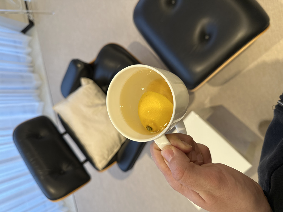 A hand holds a white mug filled with a yellowish liquid and two lemons. Behind the mug is a white rectangular object. The background includes two black, mid-century modern style lounge chairs, one with a cream-colored pillow. The setting appears to be a modern room with a window covered by blinds.