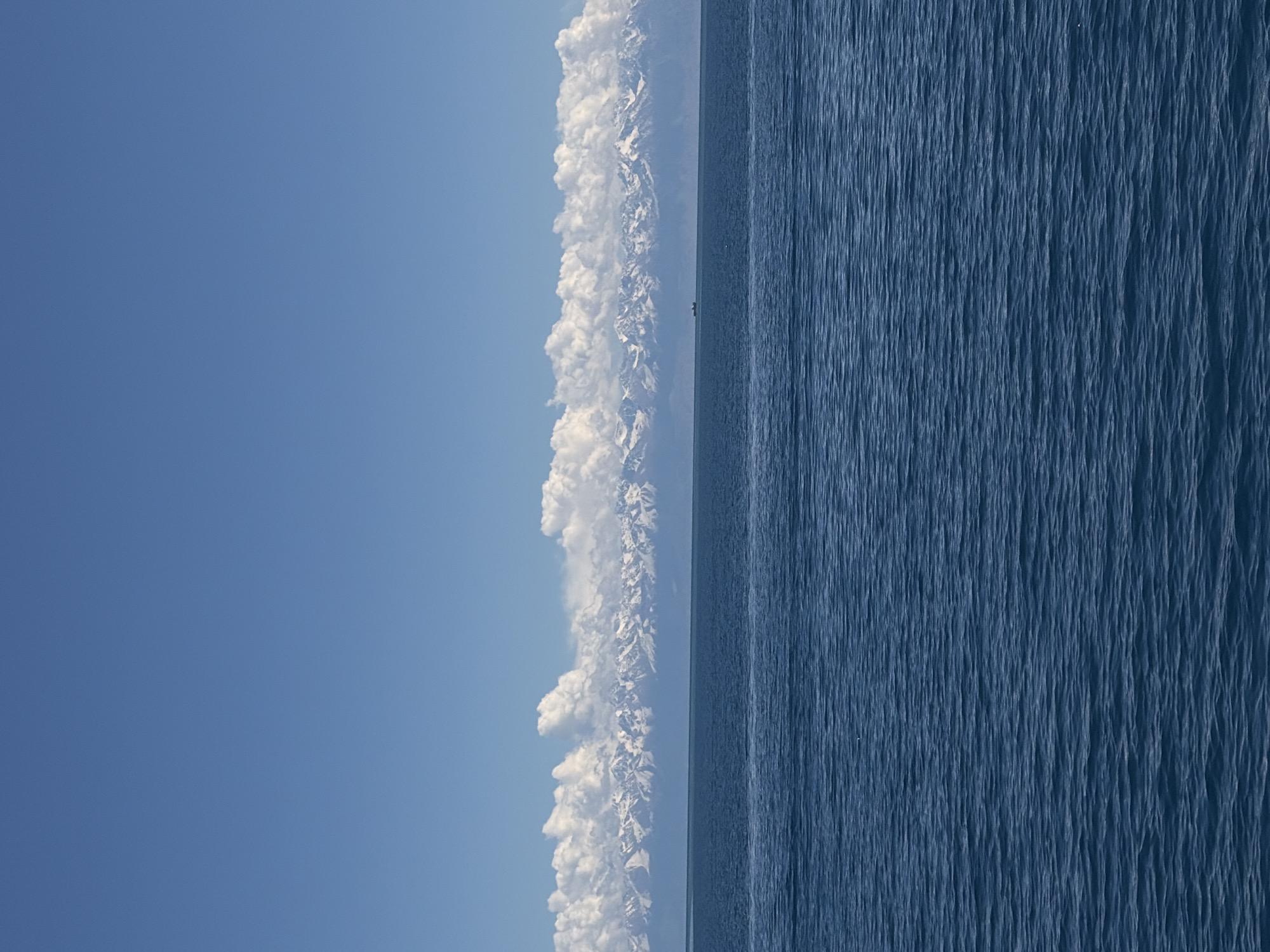 The image shows a landscape with a blue sky on the left, white clouds and snow-covered mountains in the middle, and a blue body of water on the right. The scene is oriented vertically. The location may be near Çolpon-Ata, Kyrgyzstan, due to the presence of Lake Issyk-Kul and the Tian Shan mountains in that area.