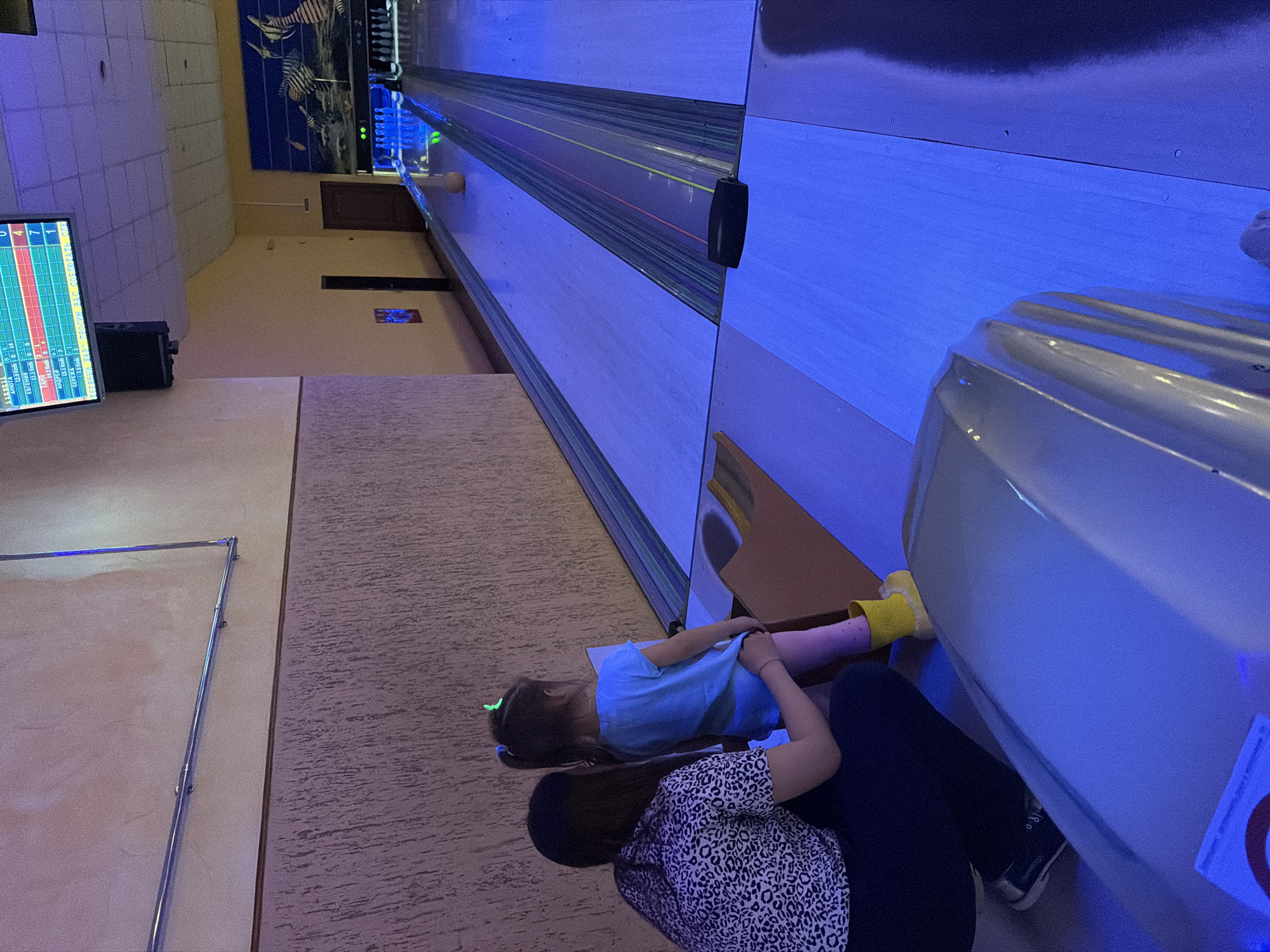 Here is a detailed description of the image:
The image shows a bowling alley, potentially in the city of Chaikovsky, Russia, given the text displayed on the score screen. A child in a blue shirt, pink leggings, and yellow bowling shoes sits near the ball return, seemingly being assisted by an adult woman wearing a spotted shirt and dark pants. A bowling lane extends into the background. The lanes are illuminated with a blue light, casting a strong hue over the scene. A score screen with Cyrillic text hangs on the wall.