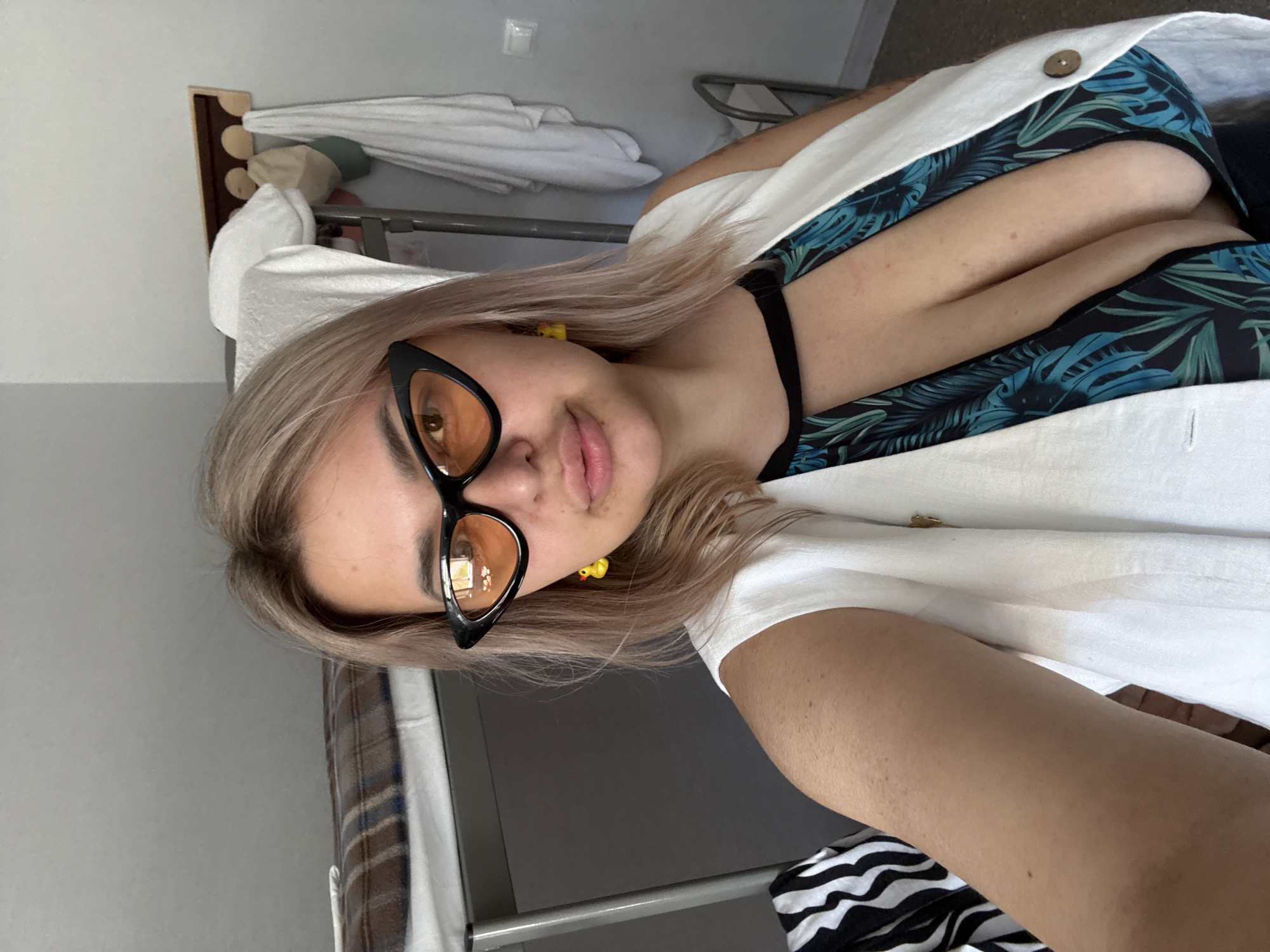 A young woman with blonde hair is taking a selfie in a room. She is wearing large black cat-eye sunglasses with orange lenses, yellow duck earrings, a white sleeveless vest over a tropical-print top with a plunging neckline, and dark bottoms. The woman is looking at the camera with her mouth slightly open. Behind her, a bunk bed with a patterned blanket is visible. To the right of the bunk bed, a wooden rack with three hooks holds two hats and two white towels. A metal railing is also present in the background. The room appears to be a simple, possibly shared living space, with plain walls. The overall lighting suggests it is daytime. The scene is consistent with a private residence, a dormitory, or a budget accommodation. The specific location in Birobidzhan, Russia, cannot be determined from the visual information alone.