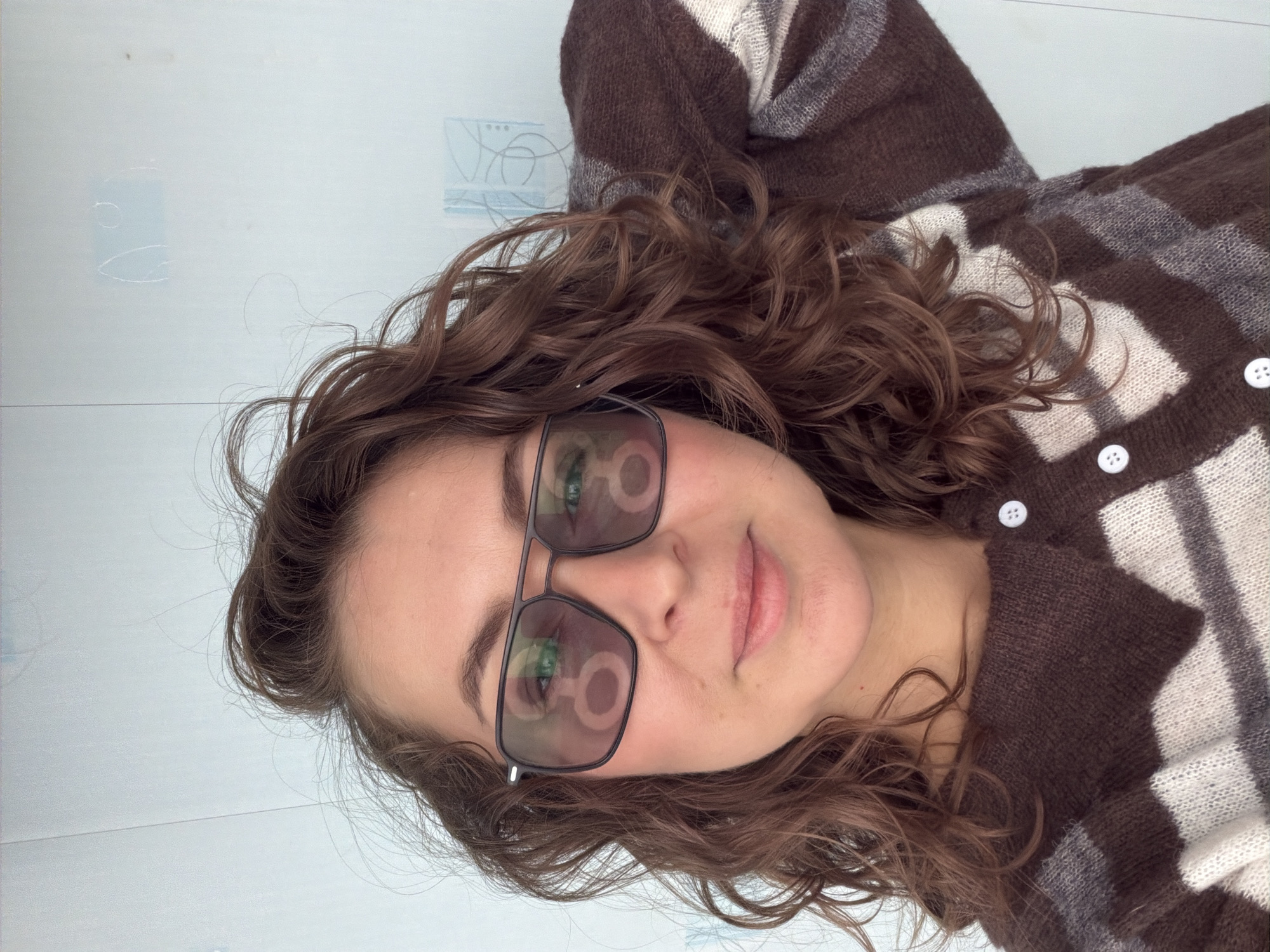 A person is lying on a light-colored, possibly white, surface. They are wearing a brown and white striped cardigan, and dark-framed glasses with lens reflections. Their hair is curly and brown. The location appears to be indoors, on a flat surface. No specific details indicate it is in Unknown, Unknown.