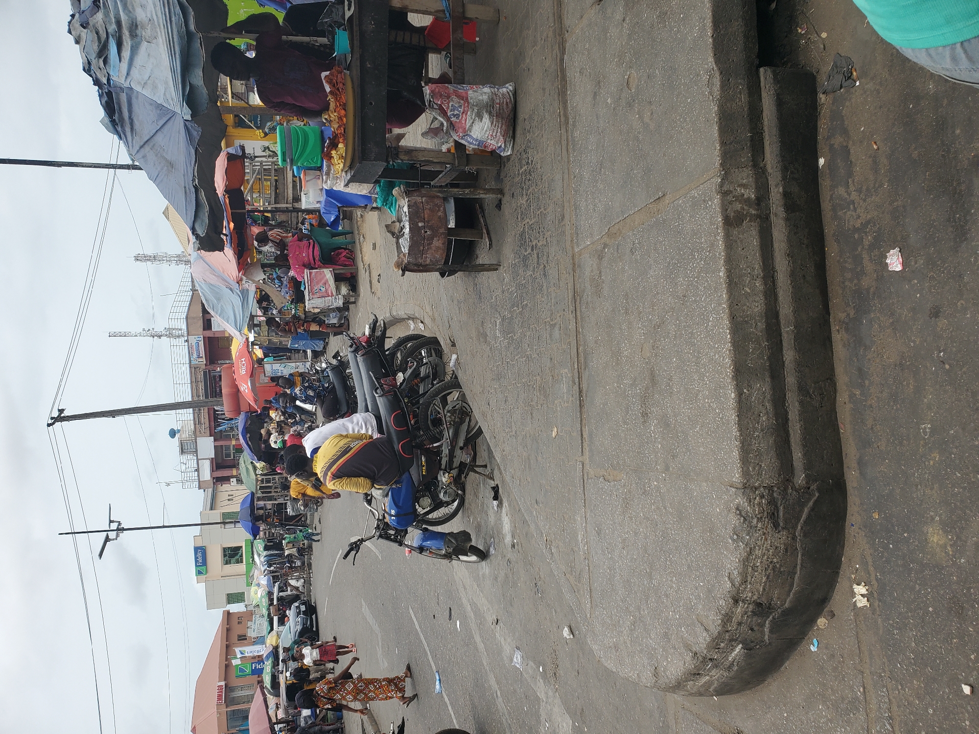 This media file captures a bustling street scene, likely a market or commercial district, in an urban environment, consistent with Lagos, Nigeria. The image is oriented vertically, showing a paved street and a raised concrete sidewalk or curb on the right.
Numerous people are visible, engaging in various daily activities. Some individuals are walking on the street and sidewalk, while others are gathered around market stalls. Several vendors can be seen behind makeshift counters, some covered by tarpaulins or umbrellas. One vendor appears to be selling fried snacks from a tray. Many motorcycles, commonly known as okadas, are present; some are parked, and others have riders, suggesting they are either waiting for passengers or are in transit. One person in a yellow and black top is seated on a motorcycle.
The setting is characterized by a row of buildings lining the street in the background, interspersed with utility poles carrying numerous power lines. The sky is overcast, indicating a cloudy daytime. The ground, both on the street and sidewalk, shows signs of wear and contains scattered litter, typical of a busy urban area.
Notable details include the market stalls fashioned from various materials, displaying goods that are mostly indistinct, though one visible bag clearly reads "CEMENT." Several umbrellas and awnings provide shade for the stalls. Visible text includes multiple "Fidelity" bank signs on buildings along the street, a red awning with "RICH" written on it, and the word "SOL" on a blue motorcycle. Another building appears to have "EMBAGO" written on it. The overall impression is one of active commerce and daily life in a vibrant city.