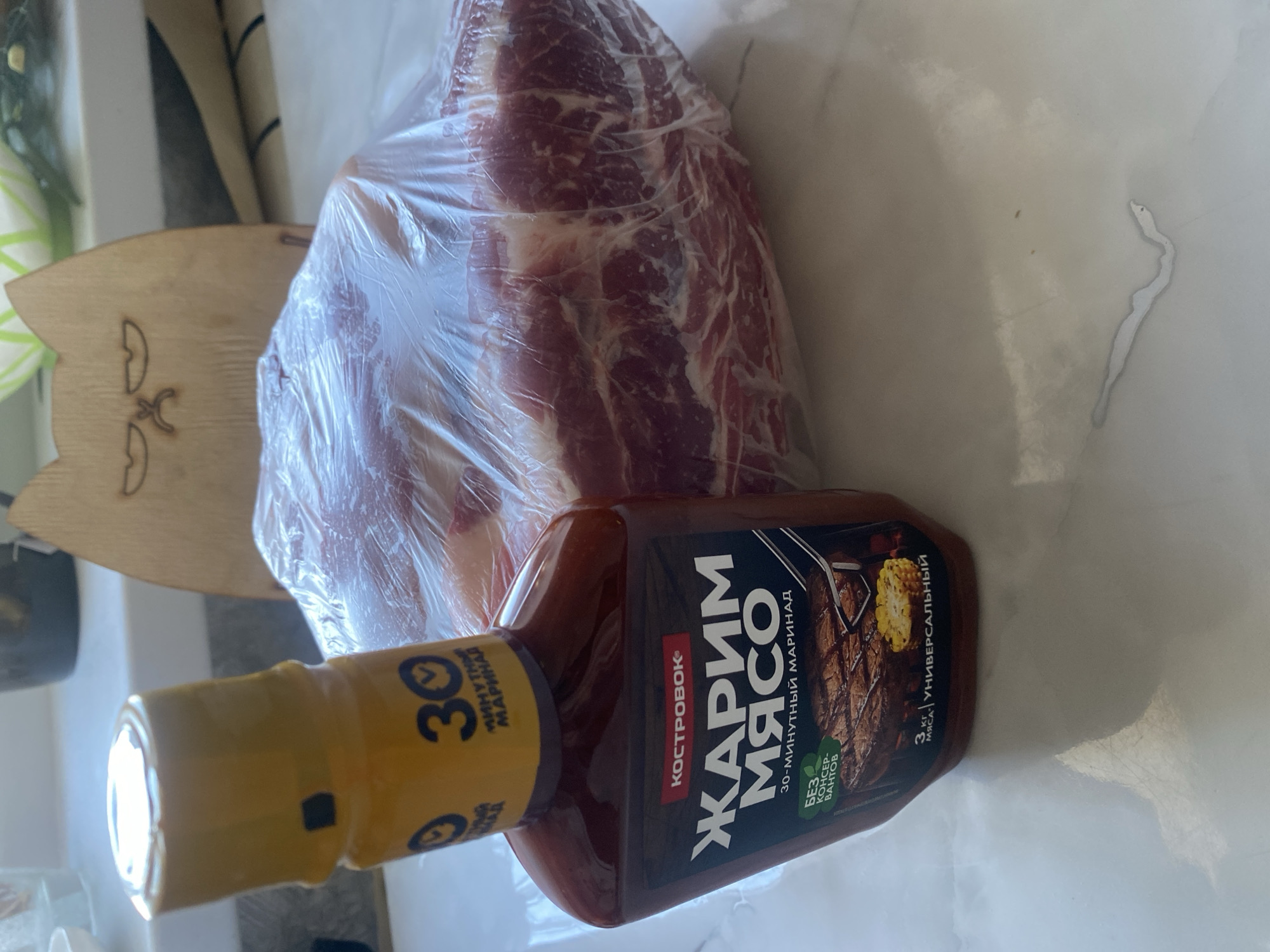 A bottle of "Zharing Myaso" 30-minute marinade stands next to a large piece of raw meat wrapped in plastic. The meat is marbled with fat and appears to be pork. The marinade bottle is dark red with a yellow cap and features text in Russian, including "Zharing Myaso," which translates to "Frying Meat." The label also indicates "30-minute marinade" and "without preservatives." A wooden cutting board with a cat-like design is visible in the background. The items are placed on a white countertop. The scene is indoors, likely a kitchen. There is no information to indicate the location is in the city of Mamontovo, Russia.
