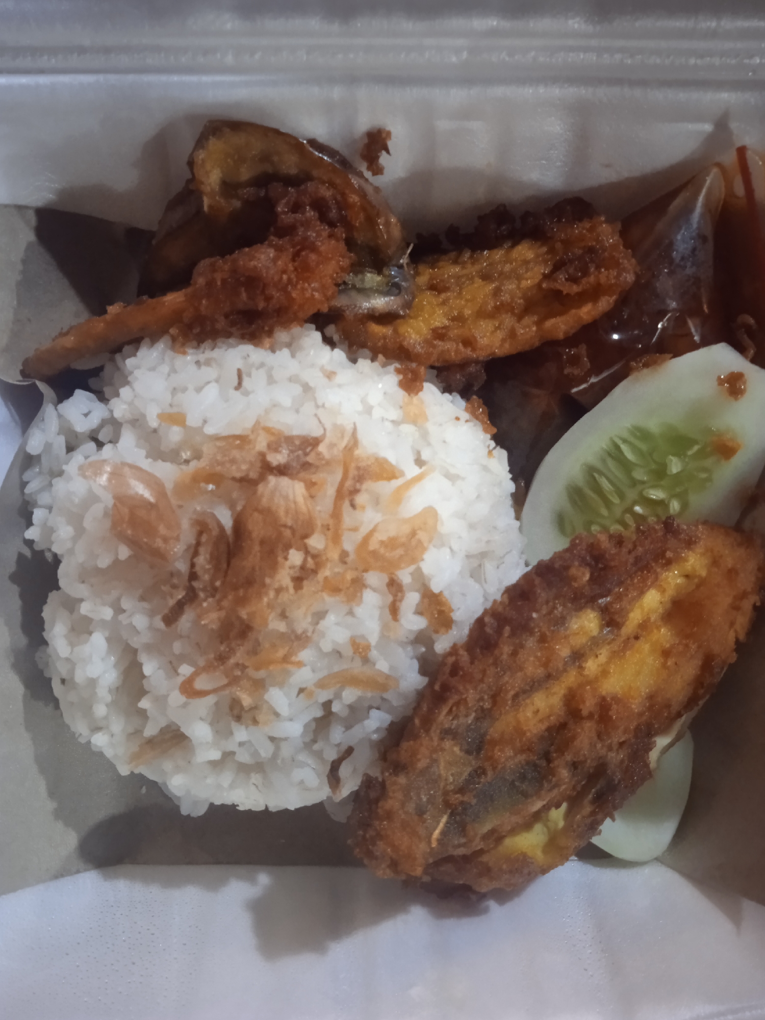 A takeout meal is depicted from a top-down perspective within a white, possibly styrofoam or paperboard, container. A mound of white rice, generously topped with golden-brown fried shallots, occupies the central-left area. To the left and above the rice, a slender, dark golden-brown fried item is partially visible, alongside a broader, similarly colored fried object in the upper center. A prominent, oblong piece of dark golden-brown fried food, consistent with fried fish or a fritter, is positioned in the lower right. A clear plastic packet containing a dark red sauce, likely chili or sambal, is visible in the upper right. A single slice of green cucumber, displaying seed patterns, rests on the right side of the container. The scene suggests a casual meal setting in Unknown, Unknown.