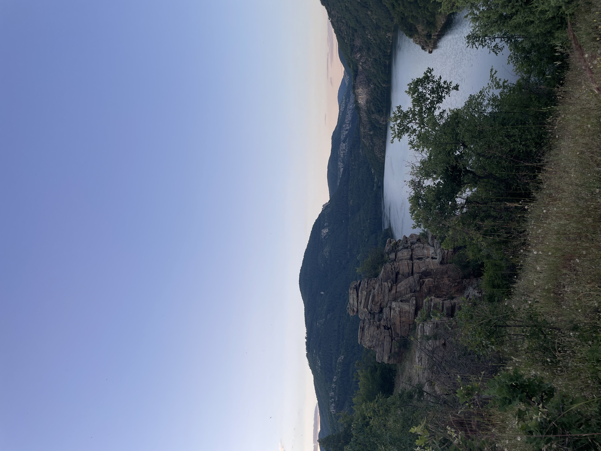 This is an outdoor, horizontal, eye-level shot of a calm, blue lake surrounded by lush green, forested hills. The sky above is a clear, pale blue, transitioning to a hint of pink and orange at the horizon where the sun appears to be setting or rising. In the foreground, on the left side of the image, is a pile of weathered, stacked rocks, appearing to be naturally formed. The terrain is covered with dry grass and scattered green bushes. The lake stretches into the distance, with hills on either side. The overall atmosphere is serene and natural. The location is identified as a viewpoint overlooking the Vacha Reservoir near Suhovo, Bulgaria. There are no people or animals visible in the image.