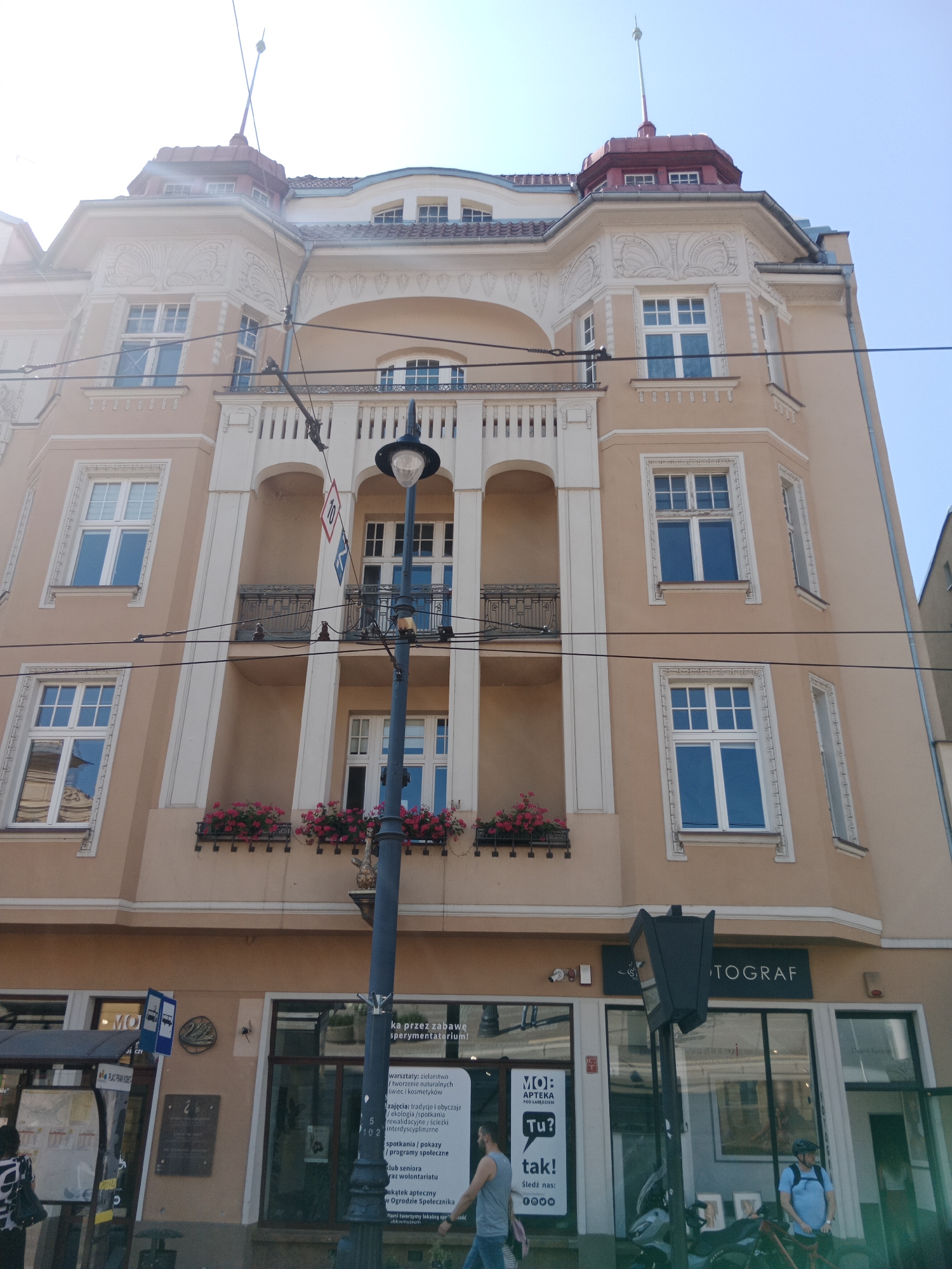 This is a street-level, slightly low-angle exterior shot of a multi-story building in Bydgoszcz, Poland. The building features a beige facade with ornate white decorative elements around windows and balconies. The upper floors have multiple windows, some with red flowers in planters beneath them. A lamppost with a globe-shaped light fixture stands in the foreground, obscuring parts of the building's lower sections.
The ground floor of the building contains storefronts. One sign reads "MOJA APTEKA POD LODOLOTEM" which translates to "MY PHARMACY UNDER THE CLOCK". Another sign above a storefront reads "-S- FOTOGRAF", likely indicating a photography studio. Several people are visible on the sidewalk. Two men are walking, one in a grey t-shirt and jeans, and another in a blue t-shirt and helmet, standing near a bicycle. A woman in a black and white patterned top is also seen on the left side of the image near a bus stop sign. The sky is clear and blue. The overall scene depicts a typical urban street environment.
