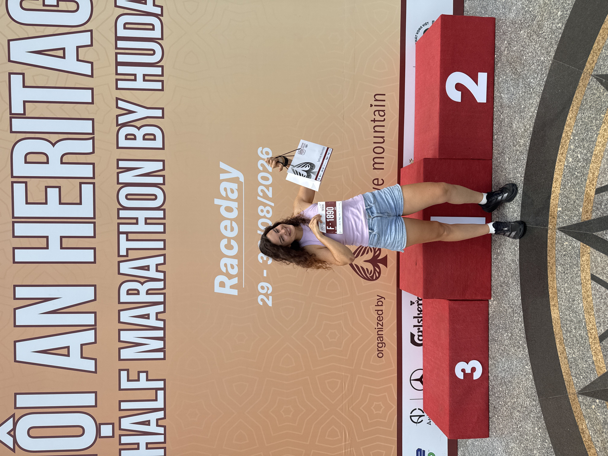 A young woman with curly brown hair stands in front of a large orange banner that reads "Hoi An Heritage Half Marathon by Huda" and "Raceday 29-30/08/2026." She is wearing a light purple tank top and denim shorts. In her right hand, she holds a race bib with the number "F-1890" and in her left, a white bag with a black handle. She is smiling and looking towards the camera.
To her right are three red podiums with the numbers "3" and "2" visible on them. The podiums are decorated with logos, including one for Carlsberg. The background banner has an intricate geometric pattern. The woman is standing on a textured grey floor with a circular pattern. The image appears to be taken outdoors in Hoi An, Vietnam, likely at the race venue.