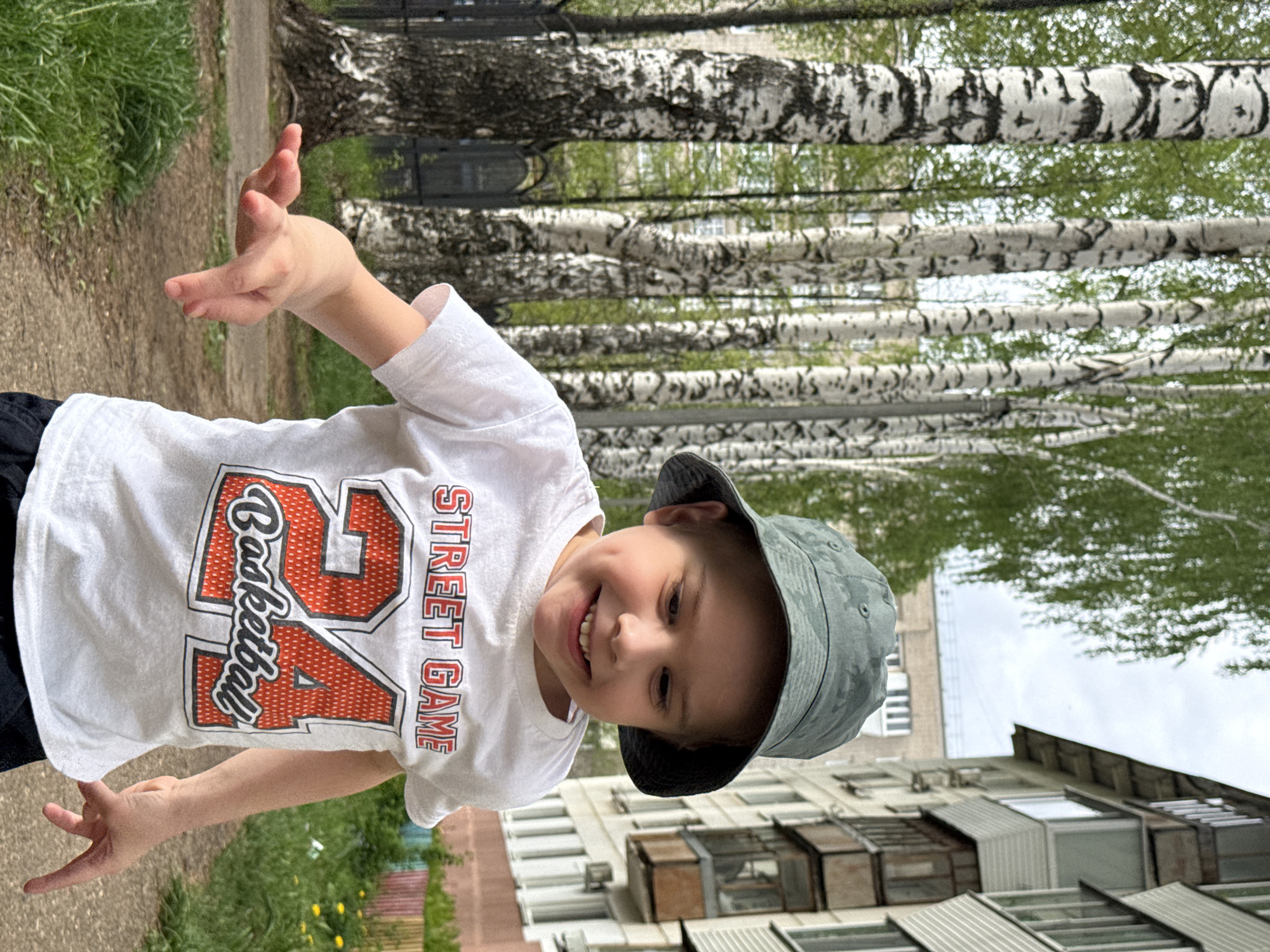 A young child is pictured outdoors in Chaikovsky, Russia. The child is wearing a light-colored t-shirt with a "24 Basketball Street Game" design and a green bucket hat, smiling with arms outstretched. Behind the child are several birch trees with distinctive black and white bark, and a residential building with windows and air conditioning units. There is also some greenery on the ground, indicating a park or yard setting. The photo appears to have been taken on a clear day, providing sufficient light for the scene. A path is visible on the left side of the photo.