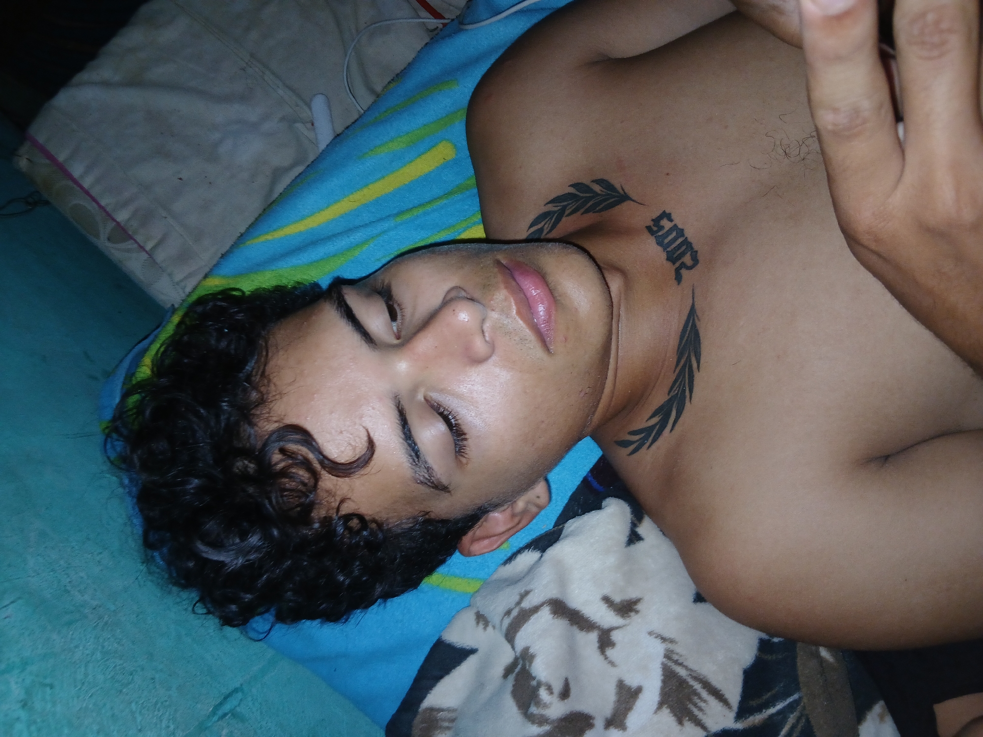 A young man with curly black hair and a tattoo on his neck of the year "2002" surrounded by leafy branches, is lying on a blue and yellow striped blanket and a patterned pillow. His eyes are closed. A white pillow is visible in the background. The image's location in Altagracia, Venezuela, is not discernible from the visual information present.