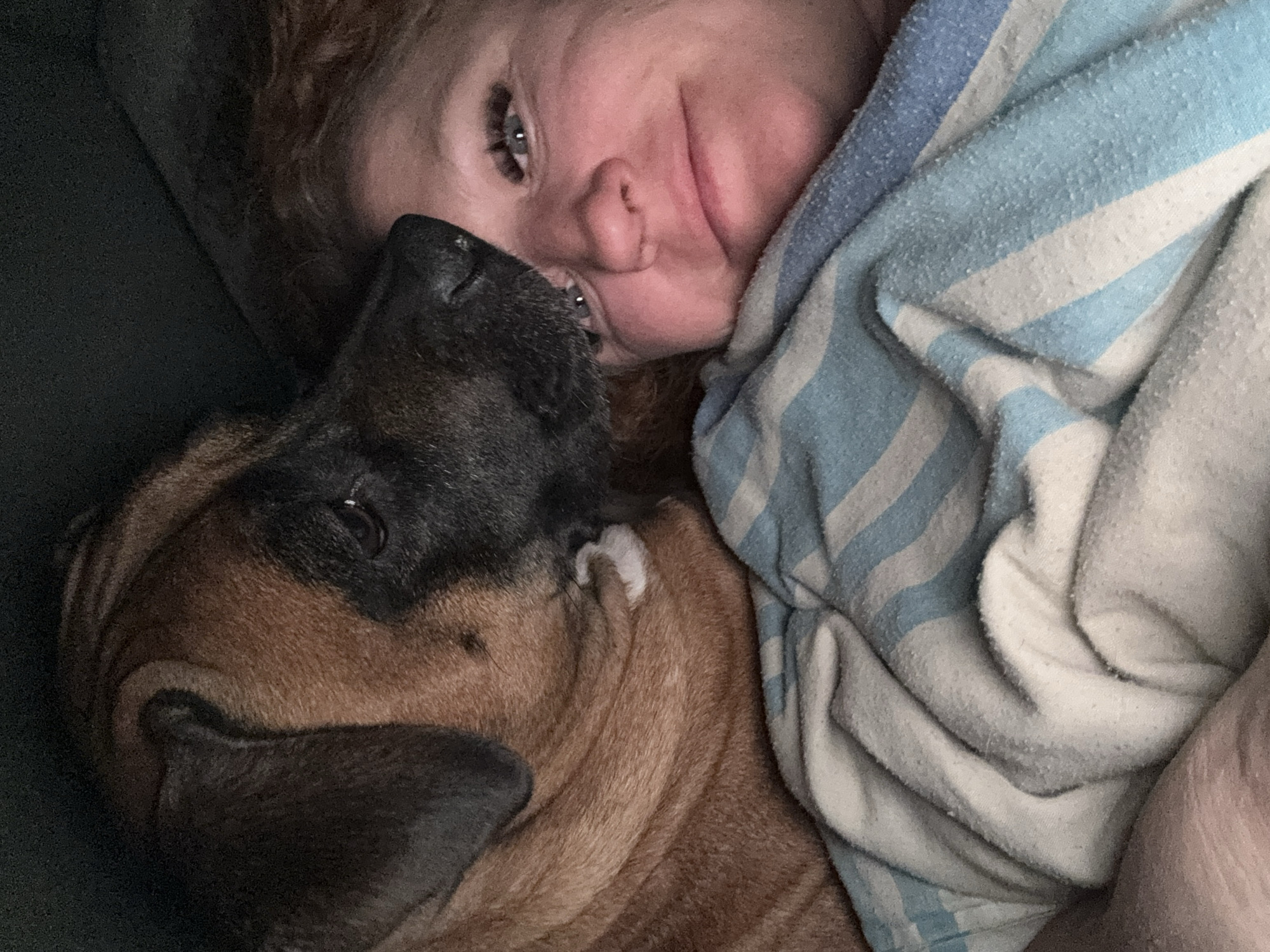 A woman with reddish-brown hair lies on her side next to a brown and black dog. The woman's face is visible, and she appears to be smiling slightly. A blue and white striped blanket is draped over her body. The dog is lying close to her, with its head near her face. The scene seems to be indoors, possibly in a home. There is no visual information to determine if the image was taken in the city of Chaikovsky, Russia.