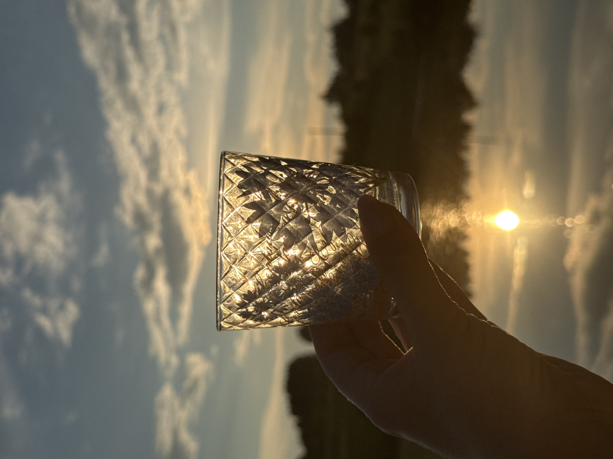 A hand holds a clear glass tumbler with a diamond pattern to the light. The glass is partially filled with an unseen liquid and reflects the setting sun. The background shows a body of water, a tree line on a distant shore, and a sky with scattered clouds illuminated by the golden hour light. The scene is likely outdoors, possibly near a lake or river, in Вассята, Russia. The sun is low on the horizon, casting a warm glow and creating a lens flare effect on the water. The image is framed in a way that emphasizes the interplay of light and shadow through the textured glass.
