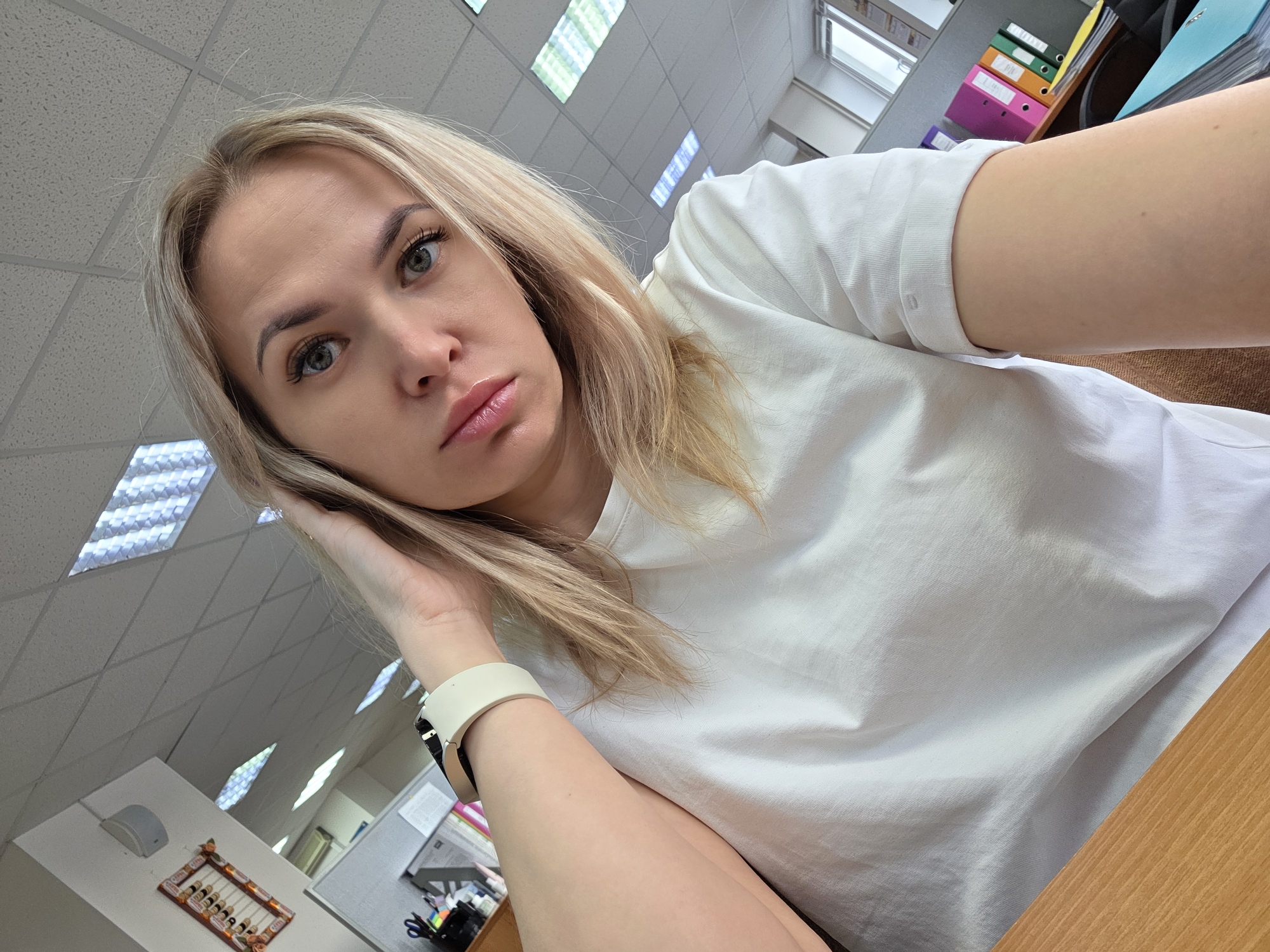 A blonde woman is taking a selfie in an office setting, likely indoors in the city of Chaikovsky, Russia. The woman is wearing a white t-shirt and has a smartwatch on her left wrist. She is resting her hand on her head, looking at the camera with a slight frown. The background consists of an office ceiling with fluorescent lights, a window, and office supplies, including files and folders. A desk is partially visible in the bottom right of the frame. An abacus, supplies and furniture are visible in the background.