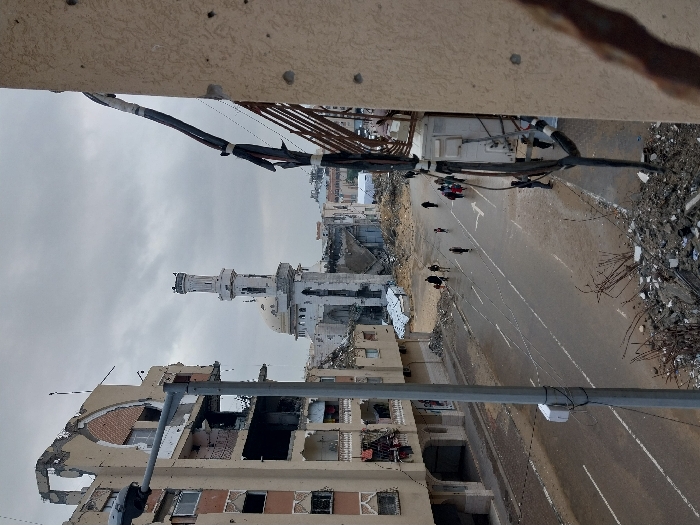 The image presents an urban landscape in Khan Yunis, Palestinian Territory, characterized by extensive structural damage under an overcast sky.
In the foreground, a pile of concrete rubble and twisted rebar lies on the ground, adjacent to the frame's bottom right. A beige building facade on the right edge displays multiple circular impact holes. Bundled electrical cables are affixed vertically to this facade. On the left, a grey lamppost stands upright.
The midground features a wet asphalt road, appearing to be a main thoroughfare, with faded white lane markings including an arrow pointing right. Numerous pedestrians, consisting of individuals and small groups, walk along both sides of the road. One group on the right side of the road includes approximately ten people, both adults and children, walking in the same direction. Another smaller group walks on the left side.
In the background, a multi-story building on the left exhibits severe damage, with large sections of its outer walls demolished, exposing interior rooms and structural elements. Arched window openings are visible on higher floors. Further back and slightly to the right of the center, a mosque with a light-colored dome and a tall, slender minaret is significantly damaged. The mosque's dome appears intact, but its minaret shows partial collapse and visible internal structure. Debris and partially collapsed structures surround the mosque. The overall environment is one of widespread destruction and disarray.