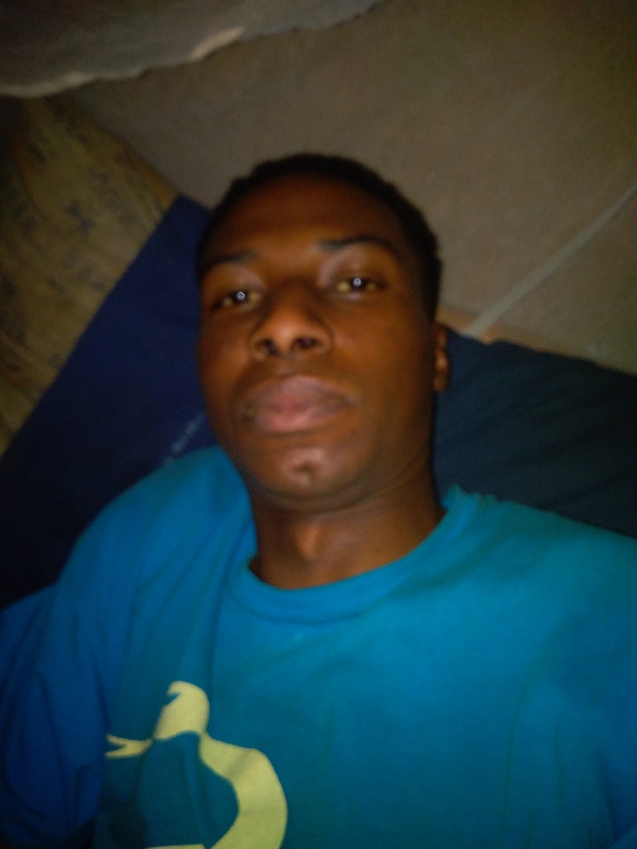 A dark-skinned man with short black hair is centered in the frame. He's wearing a bright blue t-shirt with a faded yellow graphic. The camera angle is low, looking up at the man's face, highlighting his eyes, nose, and lips. The background is dimly lit and appears to be a room with a bed or couch covered in blankets. The lighting is uneven, casting shadows on the upper part of the image.