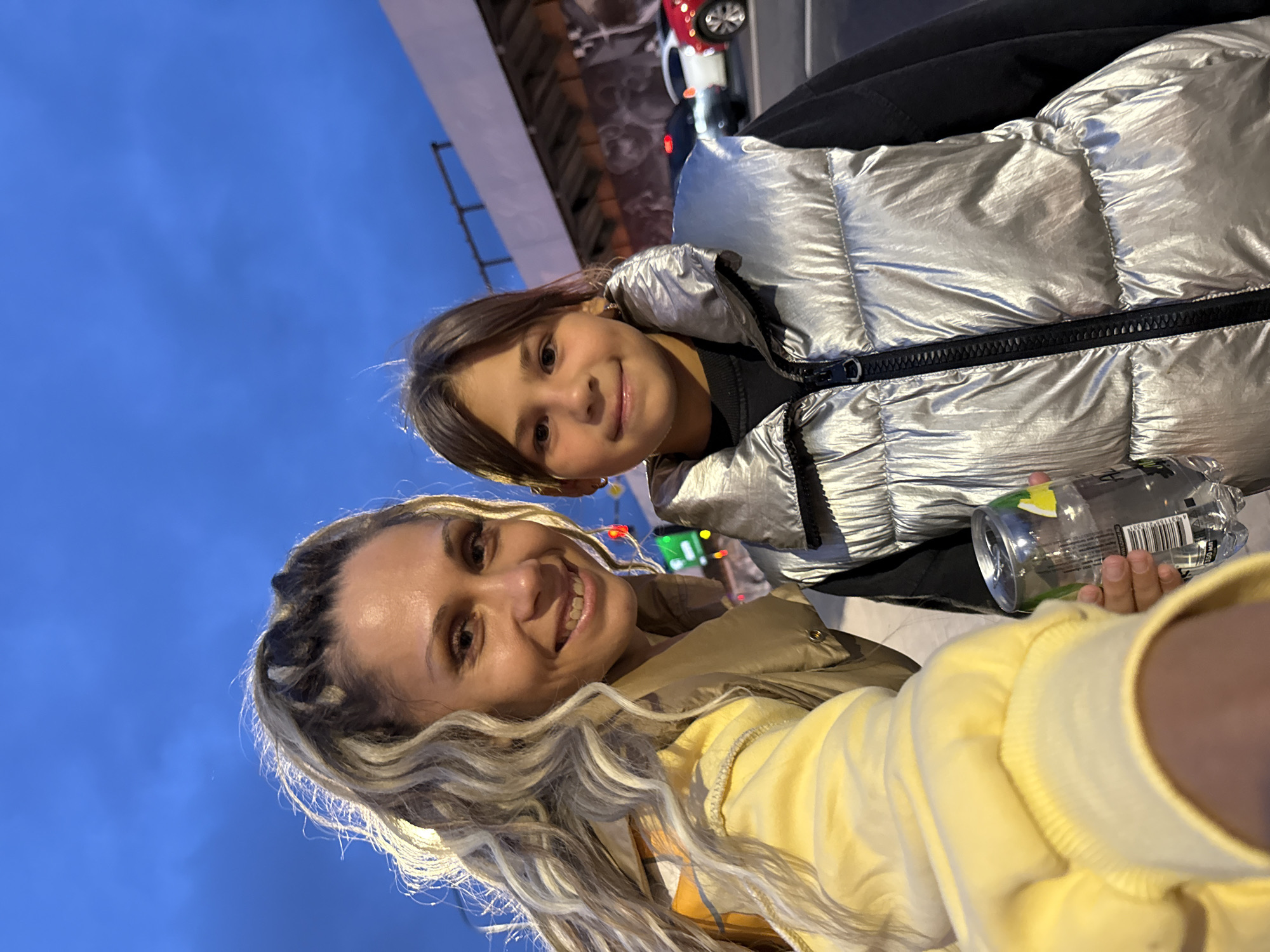 A candid selfie shows two individuals, an adult female and a young female, posing outdoors with a city skyline and cars in the background. The adult, on the left, has long blonde hair with dark streaks and is wearing a yellow jacket over a tan vest. She is smiling broadly and holding a clear plastic bottle in her right hand. The younger female, on the right, has brown hair and is wearing a silver puffer vest over a black top. She is also smiling. The sky is a deep blue, suggesting it is either early morning or late evening. The location is identified as Rusanovo, Russia.