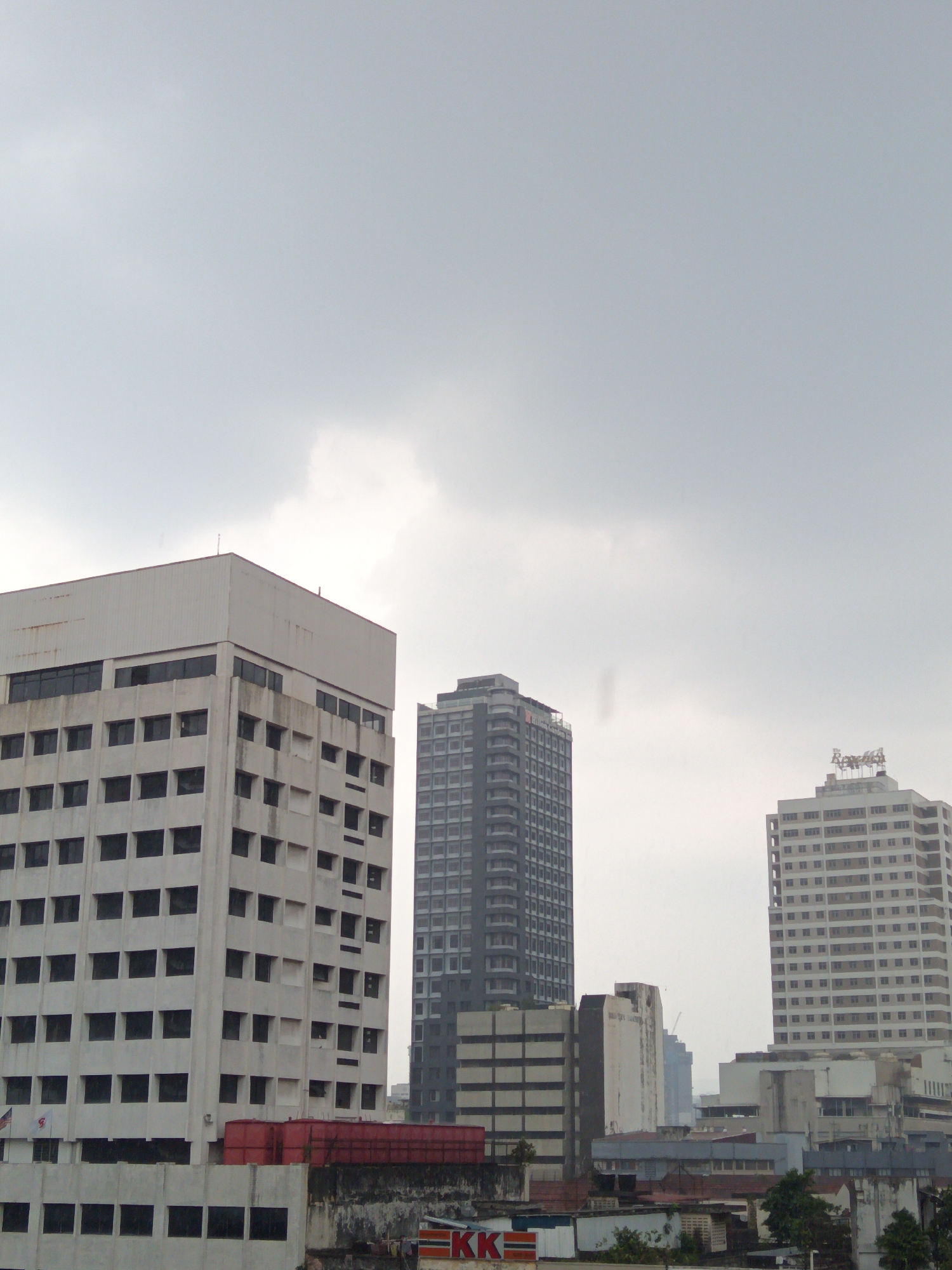 The image depicts a cityscape in Kuala Lumpur, Malaysia, under an overcast sky. In the foreground, a tall, white, rectangular building with numerous dark windows dominates the left side of the frame. To its right, a narrower, dark gray skyscraper with a repetitive pattern of windows is visible. Further to the right, another tall building, mostly white with dark windows, stands out, and atop it, a sign with red and white lettering is discernible. The lower portion of the image reveals a collection of buildings with varying architectural styles and colors, including some red structures and a wall with graffiti. The overall impression is that of an urban environment, possibly a business district, given the presence of numerous high-rise buildings.