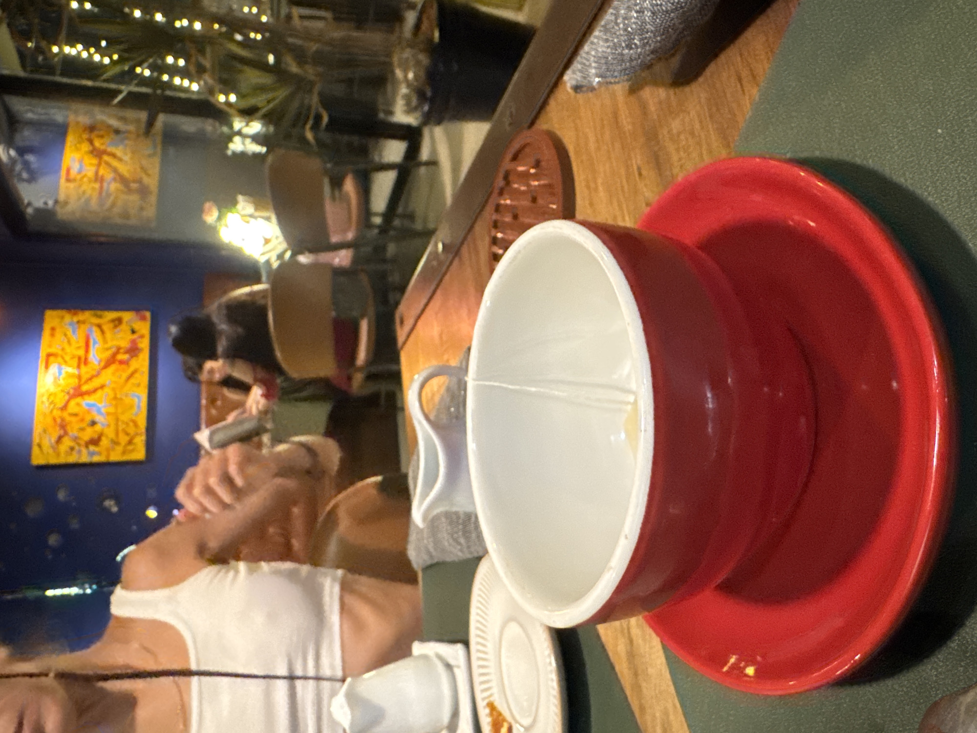 In this close-up, slightly blurry image, a person's shoulder and arm are visible on the left, wearing a white tank top. In the foreground, a red bowl rests on a red saucer, both on a dark green placemat. Inside the bowl, a clear plastic utensil is submerged in a light-colored liquid. To the left of the bowl, a small white pitcher sits on a white square coaster, and further left, a small white plate with a textured rim holds a tiny amount of orange food. Behind these items, a grey cloth is draped. The background shows other patrons sitting at tables in a dimly lit restaurant with blue walls adorned with colorful abstract art and string lights. The overall scene suggests a meal or drink service in an indoor establishment, possibly a restaurant or cafe, in Hanoi, Vietnam.
