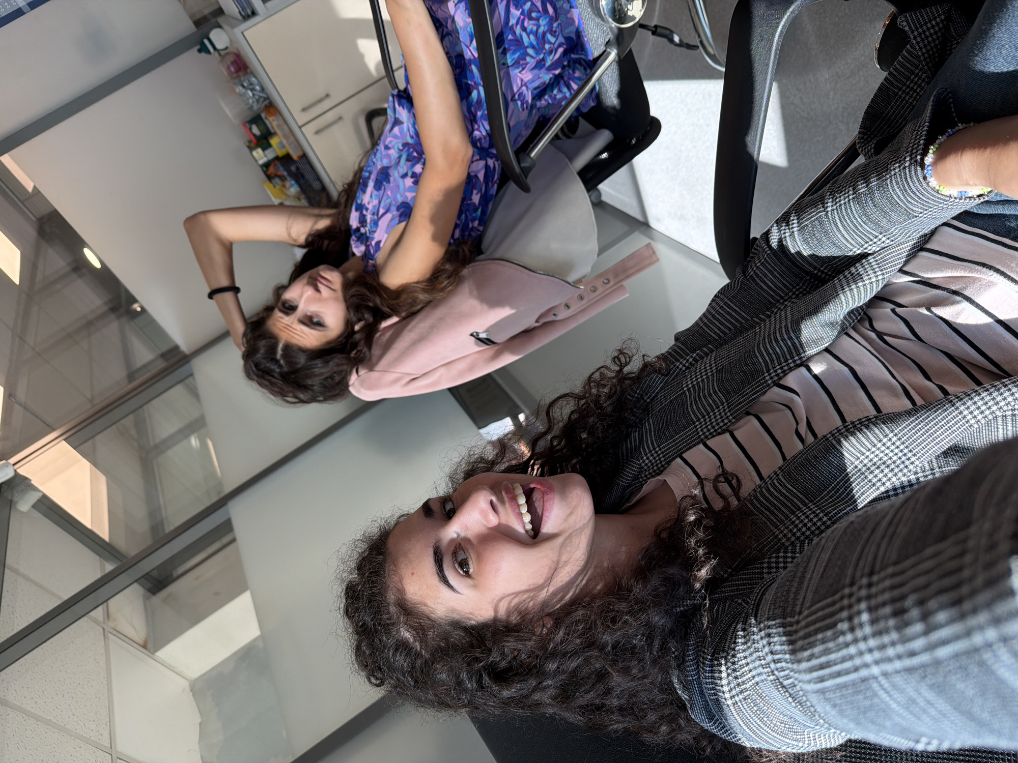 Two young women are seated in office chairs, possibly in a modern office setting within Sofia, Bulgaria. The woman in the foreground, on the left, is closer to the camera and smiling broadly, looking directly at the viewer. She has dark, curly hair and is wearing a grey and white checkered blazer over a striped t-shirt. Her left arm is resting on her lap, and a colorful beaded bracelet is visible on her wrist.
The second woman, in the background and to the right, is leaning back in her chair with her arm raised and hand behind her head. She has long, dark hair and is wearing a purple floral-print dress and a light pink jacket draped over the back of her chair. Her expression is more relaxed and perhaps a bit playful or weary.
The environment appears to be an office with large windows reflecting light. Desks or cabinets are visible in the background, along with some office supplies. The lighting suggests it might be daytime, with sunlight casting shadows on the floor and furniture. The overall impression is informal, perhaps a break or casual moment during a workday.