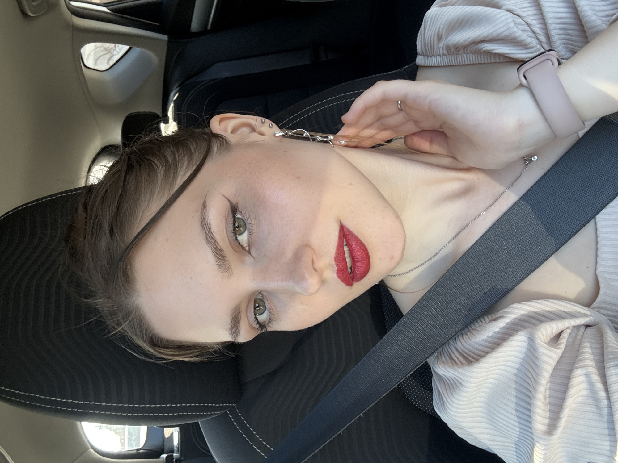 A young adult female is depicted in a close-up portrait shot from within a vehicle. She has light skin with visible freckles, green eyes enhanced with black winged eyeliner and mascara, and wears vibrant red lipstick on her slightly parted lips. Her dark brown hair is styled back, with a few loose strands framing her face. She wears a light-colored, possibly pale pink or beige, ribbed top with puffed sleeves. A silver chain necklace with a small clear pendant is visible, along with a silver ring on her left ring finger and a light pink smartwatch on her left wrist. Her left ear features multiple piercings, including two small studs and a long, elaborate dangle earring composed of silver-toned elements and clear adornments. Her left hand is positioned with fingers gently resting against her neck/jawline. She is secured by a dark grey textured seatbelt, running diagonally across her chest. The background consists of a dark fabric car seat with white stitching, visible sections of the car's interior ceiling, and windows that show blurry outdoor elements. Natural light illuminates the scene from the right. No specific visual cues are present in the image to definitively identify the location as Saint Petersburg, Russia, or any particular area within it.