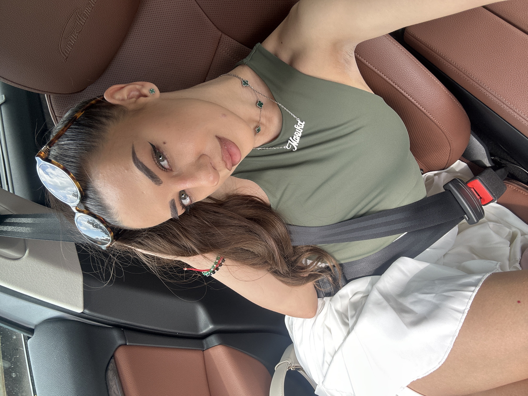A young woman with long, dark hair is lying on her side in the passenger seat of a car. She is wearing a green tank top, white shorts, and a seatbelt. She has on sunglasses perched on her head, and a necklace with clover-shaped charms and her name, "Mariska," in script. On her left ear, she has a small, green clover stud earring. She is also wearing a bracelet on her left wrist with red, green, and black beads. The car has brown leather seats with a textured pattern. The interior of the car is visible, including part of the dashboard and seatbelt buckle. The image appears to have been taken during the daytime, with natural light illuminating the scene. The woman is looking towards the camera with a relaxed expression. The background shows the interior of a car.