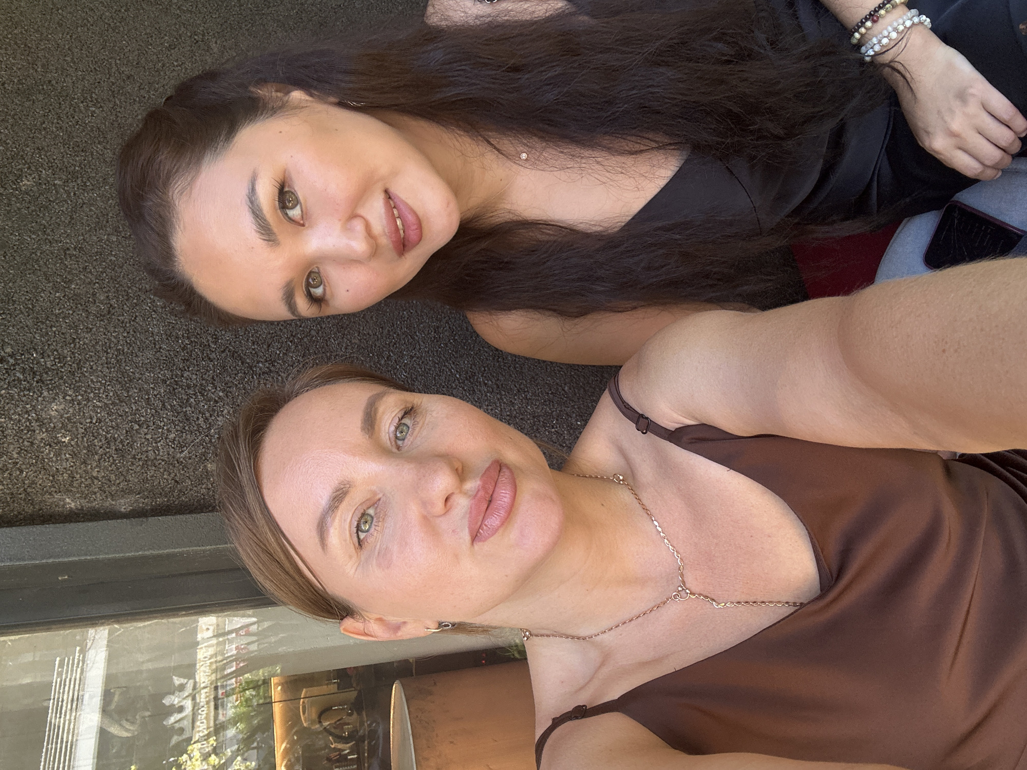 Two women are taking a selfie outdoors, appearing to be in a cafe or restaurant setting in Sochi, Russia. The woman on the left has light brown hair pulled back and is wearing a brown satin camisole and a delicate gold necklace. She is looking directly at the camera with her lips slightly parted. The woman on the right has long, dark brown hair and is wearing a black satin camisole. She is also looking towards the camera, with a slight smile. They are positioned close to each other, with the woman on the left slightly closer to the camera. Behind them, the background consists of a textured dark wall on the right and a window with reflections of trees and buildings on the left, suggesting an urban environment. The lighting indicates it is daytime.
