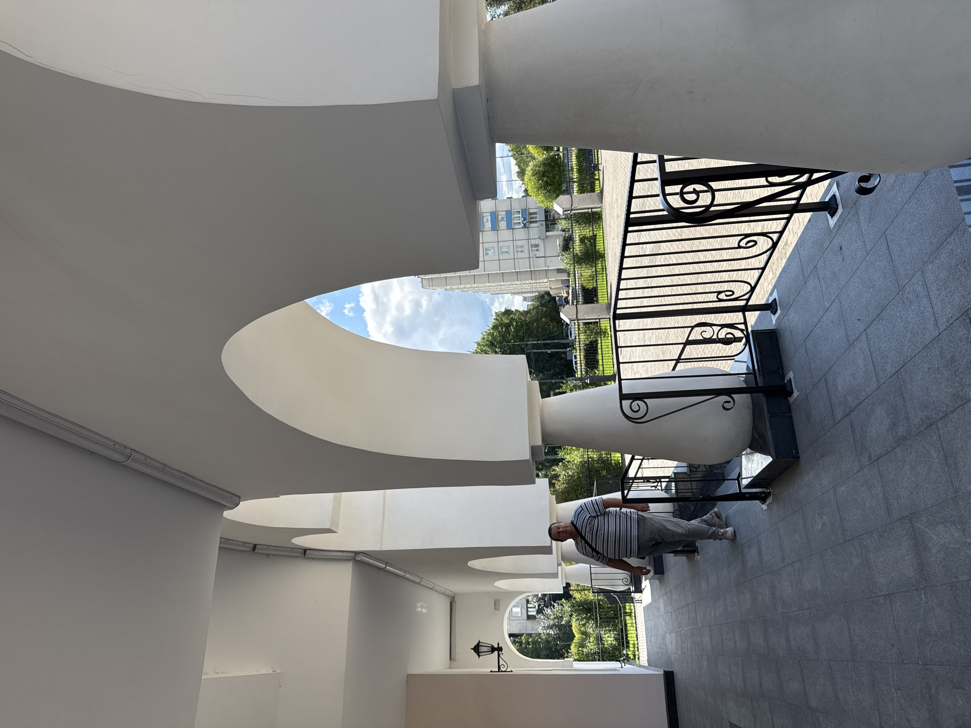 A man in a striped shirt and gray pants walks along a covered walkway with arched openings. The walkway has a tiled floor and is lined with white pillars and a black metal railing. In the background, a multi-story building and trees are visible under a blue sky with white clouds. The scene appears to be in an urban setting, possibly residential or institutional.