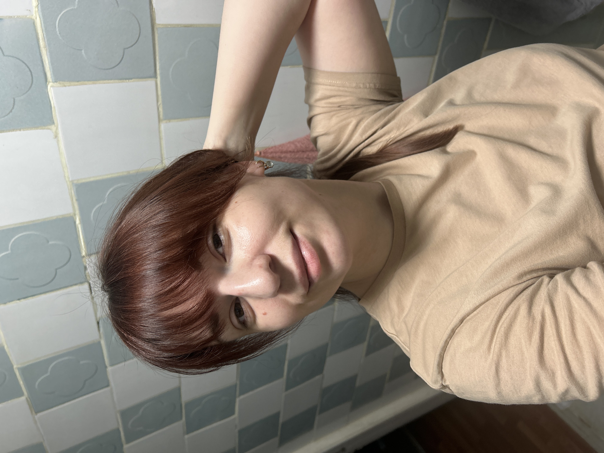 A selfie shows a woman with shoulder-length auburn hair and bangs, wearing a tan long-sleeved shirt. She is smiling slightly and is positioned against a backdrop of blue and white patterned tiles. Her right arm is raised, with her hand near her head. An earring is visible in her left ear.
The city of Фоки, Russia is not depicted.