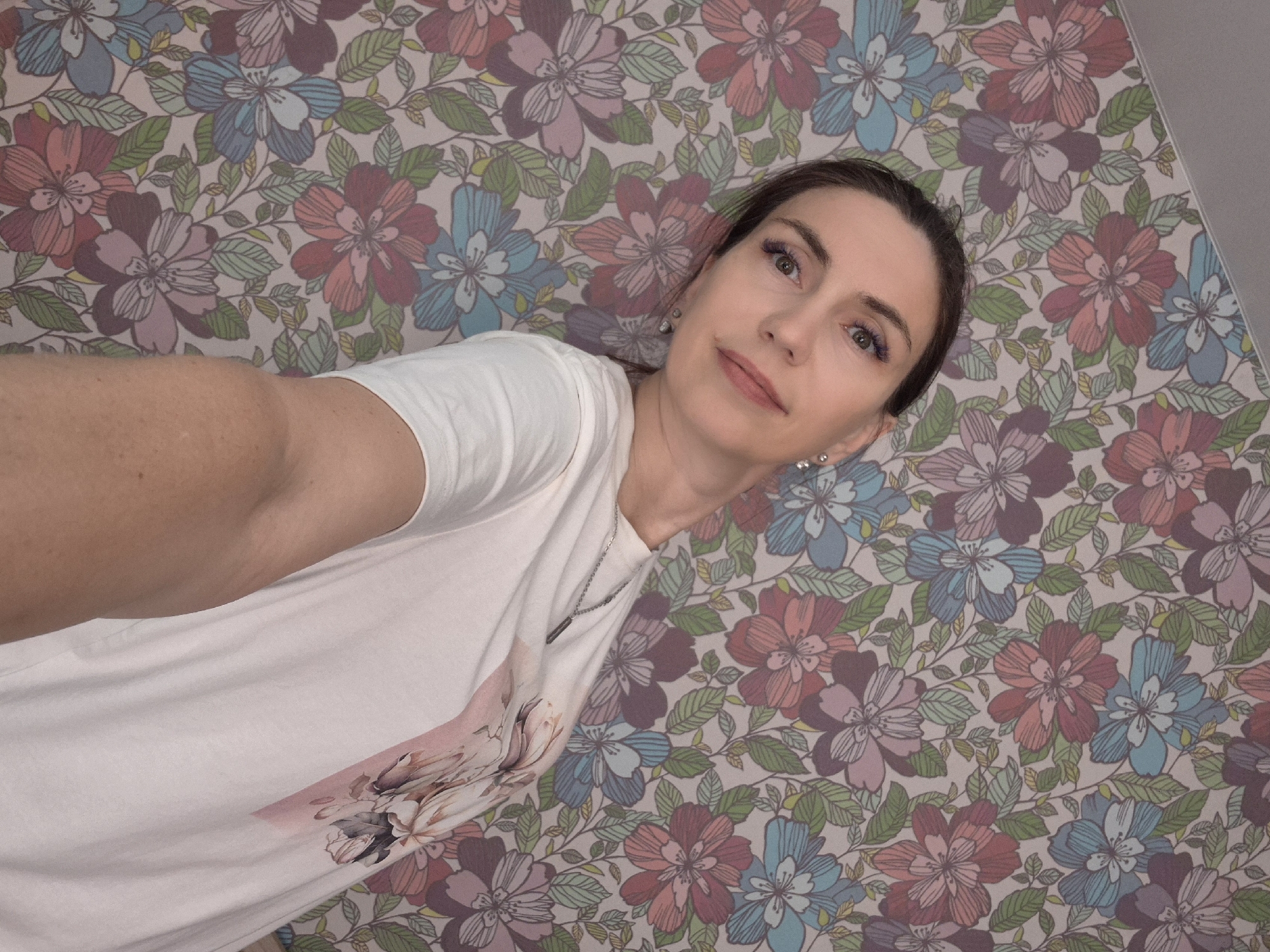 A woman is pictured from the chest up, looking towards the camera with her head tilted. She has dark hair pulled back and is wearing a white t-shirt with a floral design. Her makeup includes purple eyeshadow and lipstick. She is leaning against a wall with floral wallpaper. The wallpaper features a pattern of large flowers in shades of pink, red, blue, and purple, with green leaves and vines. There are no other people or objects clearly visible in the frame. The lighting appears to be artificial, casting a soft glow on the subject. The image was taken from a high angle, looking down at the woman. There is no visible indication of the city of Chaikovsky, Russia.