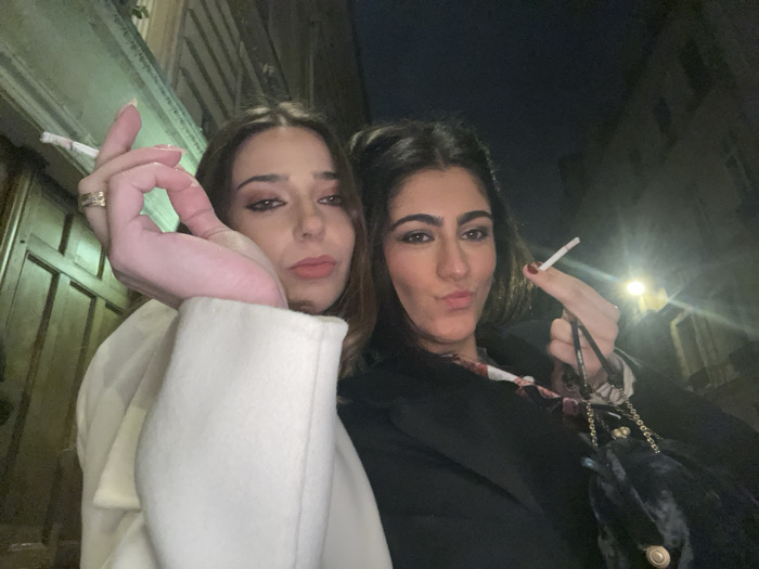 Two women are posing at night, both holding lit cigarettes. The woman on the left wears a white coat and has long brown hair. The woman on the right wears a black coat with a patterned shirt underneath and has dark brown hair. She holds a small black velvet purse with a gold chain. They are standing in front of a building with dark wooden doors and a light fixture is visible in the background.