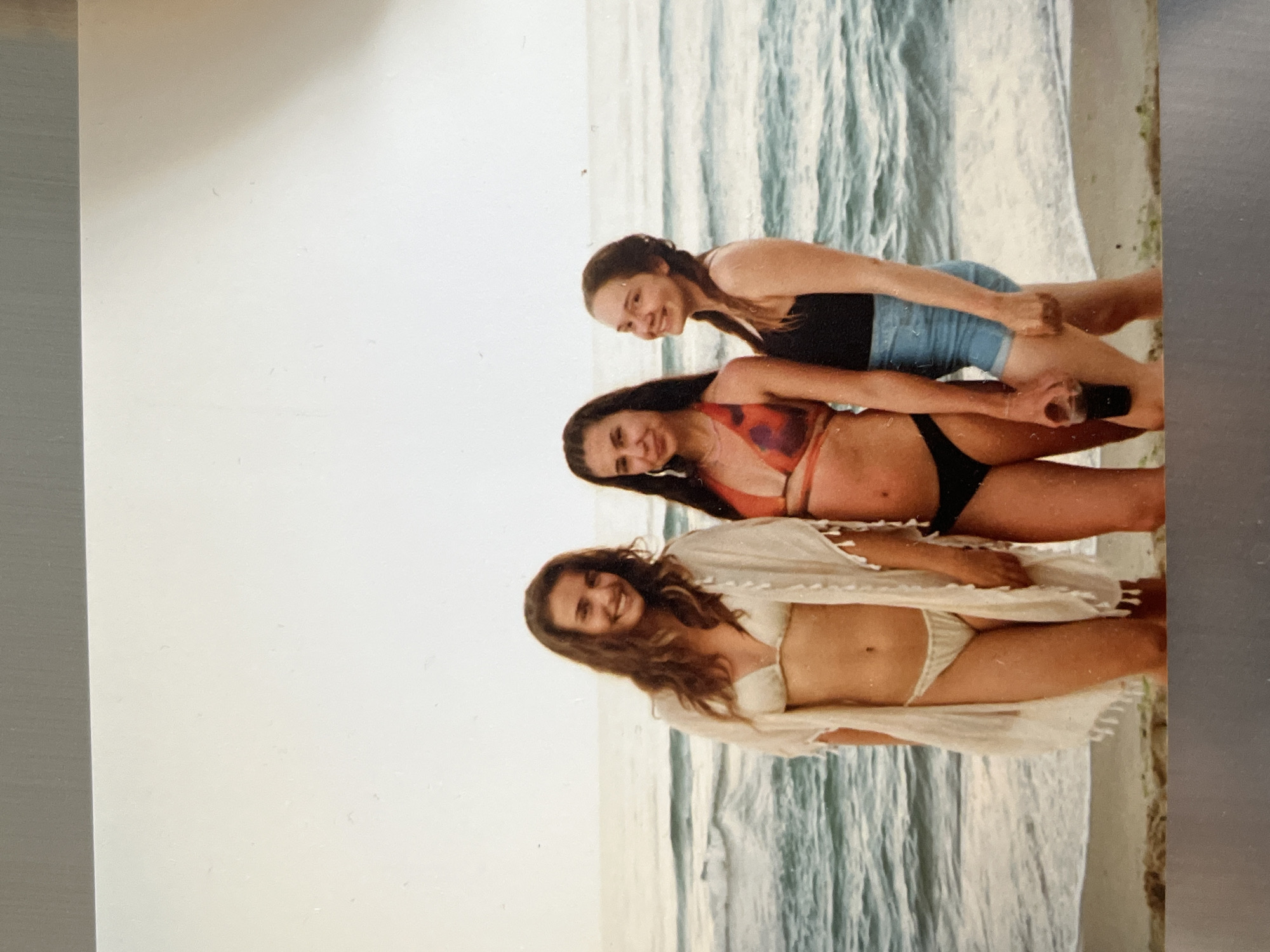 Three women stand on a beach in front of the ocean. The woman on the left is wearing a white bikini and a cream-colored cover-up. The woman in the middle is wearing an orange and black patterned bikini. The woman on the right is wearing a black tank top and denim shorts, and she is holding a dark-colored cup. The ocean waves are visible behind them, and the sky is overcast.