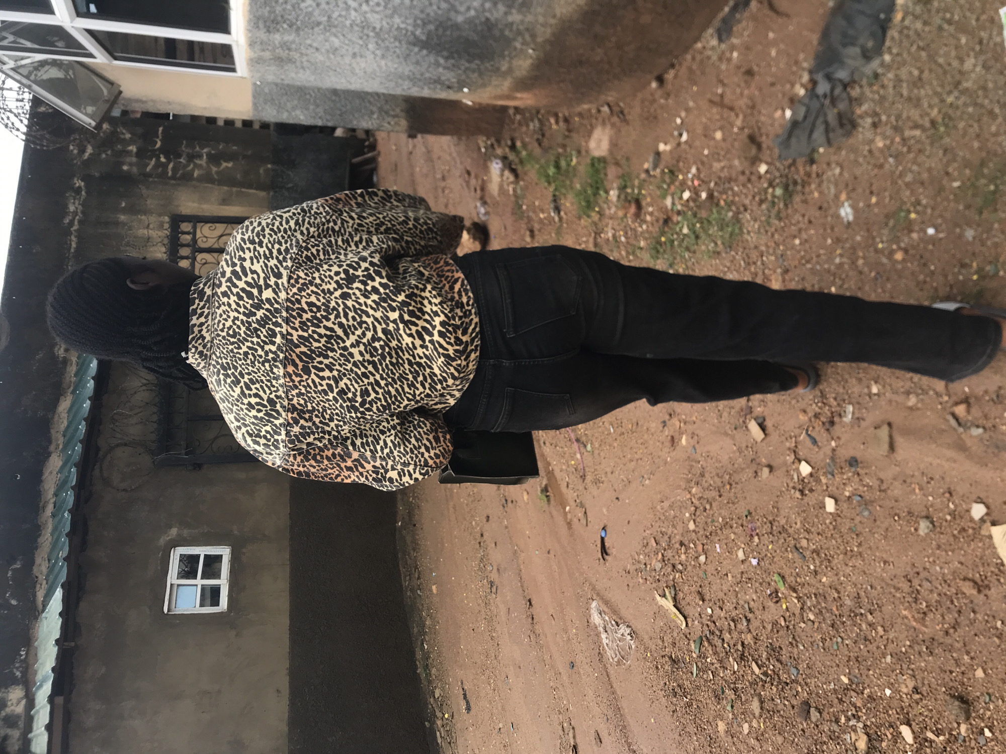 A person with braided black hair is walking away from the camera on a dirt road in Jos, Nigeria. The person is wearing a tan and black leopard print button-up shirt and black jeans. They are also carrying a black purse in their left hand. The road is lined with buildings. On the left is a building with a textured brown and grey facade. It has a small, white-framed window with four panes. A green corrugated metal roof is visible above the building. To the right of the person, a rough, grey concrete structure stands. The ground is covered in reddish-brown dirt with scattered rocks, debris, and patches of green vegetation. The sky is overcast.