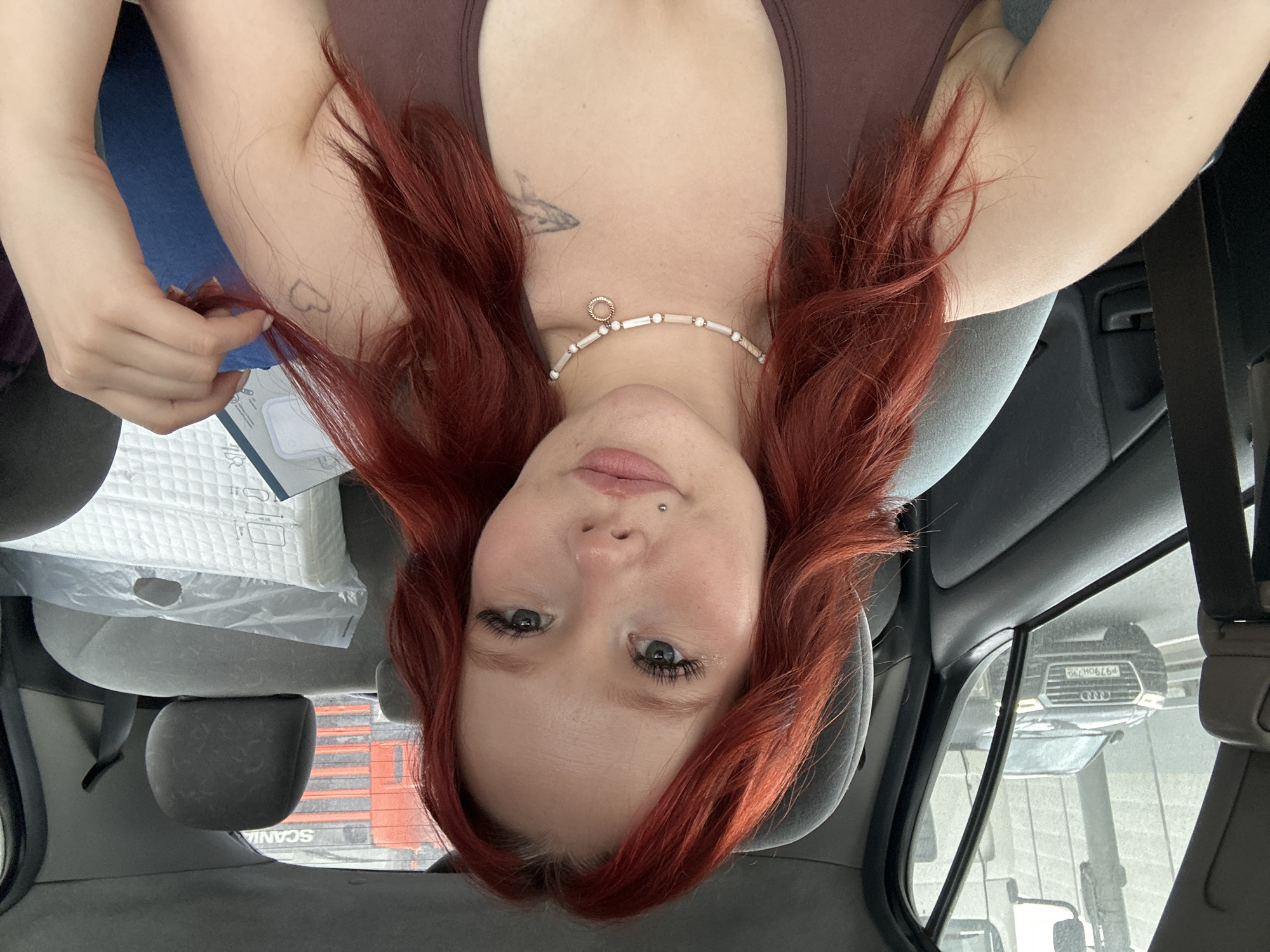 A young woman with bright red hair is sitting in the passenger seat of a car. She is looking directly at the camera, with her head slightly tilted. Her hair is styled in loose waves and falls around her shoulders. She has on a brown tank top and a pearl necklace. There is a piercing on her lower lip. In the background, through the car window, an Audi car is visible on a highway, with a large truck behind it. The interior of the car shows a gray seat and a seatbelt. There are also some items in plastic wrapping in the back seat.