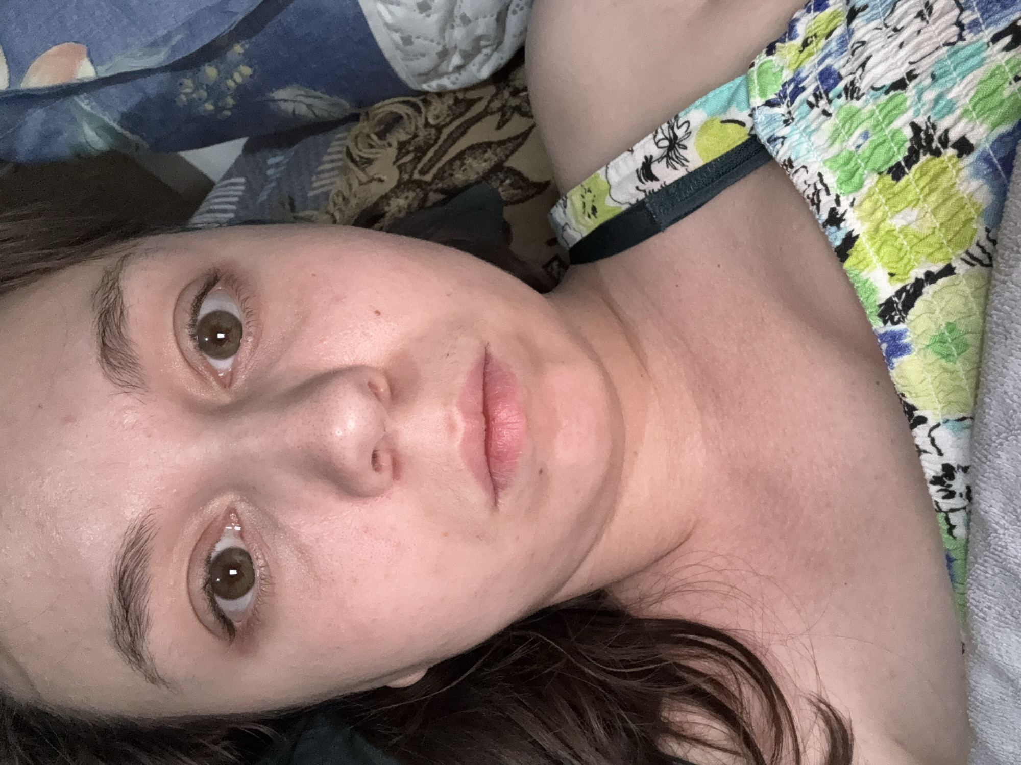A close-up, selfie-style shot features a woman lying down with her head tilted. Her brown eyes are looking directly at the camera, and her mouth is slightly open. She has light skin and brown hair. She is wearing a floral tank top with green, blue, and white colors. The background consists of bedding with a blue, patterned comforter and white lace trim. The image was taken indoors, likely in a bedroom. There is no visible information to indicate the location in Shymkent, Kazakhstan.