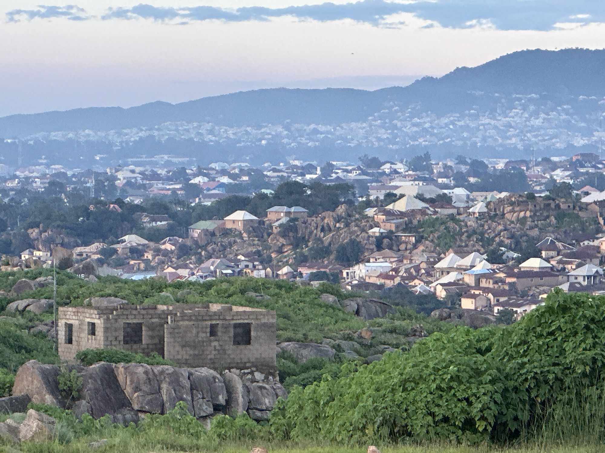 This is a wide, horizontal, eye-level shot of a sprawling, densely populated urban area in Jos, Nigeria, nestled amongst rocky hills. The foreground features a partially constructed building made of grey concrete blocks, with open window and door frames, situated on a rocky outcrop. Lush green vegetation, including grasses and bushes, surrounds the building and covers much of the rocky terrain.
In the mid-ground, the city unfolds as a dense mosaic of buildings with various roof colors, including white, grey, red, and blue. These structures are built on and around the rocky hills, creating a textured and uneven urban landscape. Trees are interspersed throughout the city, adding patches of green.
The background is dominated by a large, hazy mountain range, also covered with buildings that fade into the distance. The sky above is a soft, pale blue with wispy clouds, suggesting either dawn or dusk. The overall impression is of a vibrant, active city built into a rugged natural environment. There are no people or animals visible in the frame.