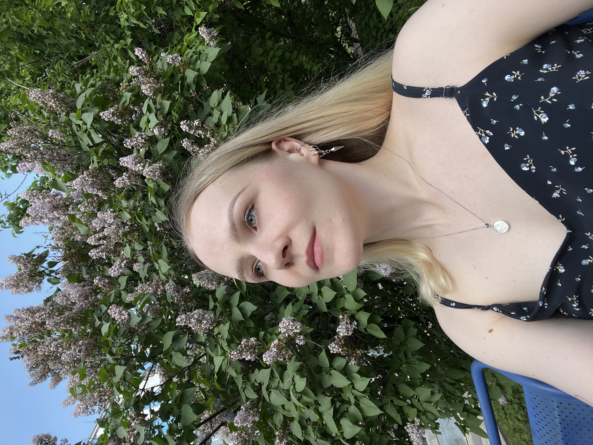 A young woman with blonde hair is positioned in front of a bush full of light purple flowers. She is wearing a black floral dress with thin straps and a silver necklace. She has multiple piercings in her left ear. The background suggests an outdoor setting, possibly a park or garden. A blue chair is visible beneath her arm. The location is not identifiable as Khimki, Russia from visual information alone.