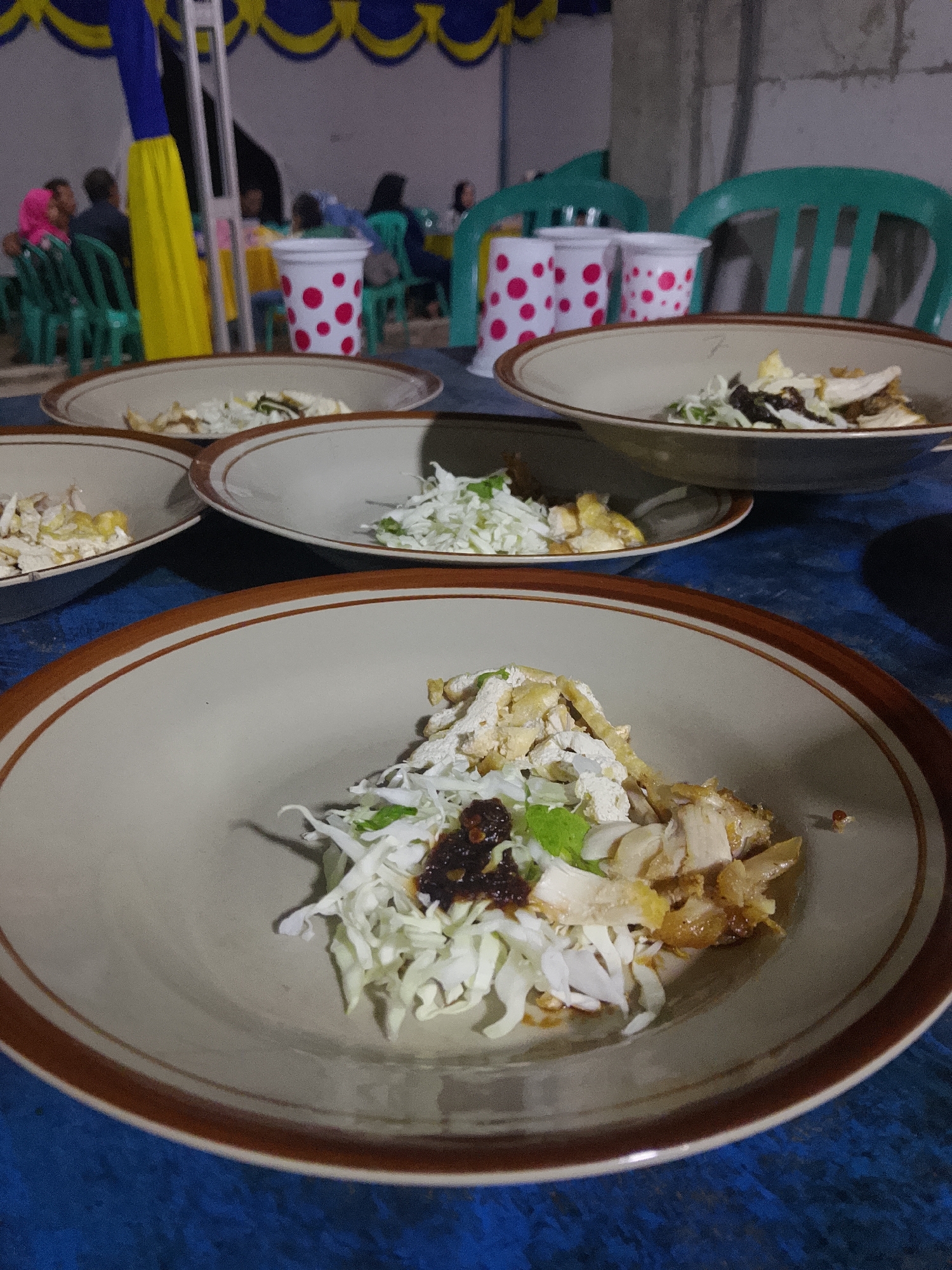 The image shows a close-up of several plates of food, likely a meal served outdoors in a casual setting. In the foreground, a large, light brown ceramic bowl with a dark brown rim is filled with shredded cabbage, pieces of chicken, and a dark, textured condiment. Behind this, other similar bowls of food are visible, arranged on a blue, textured surface. In the background, people are seated at tables, with some chairs visible, and colorful banners are hung overhead. The scene appears to be a gathering or event, possibly a meal served at an outdoor venue in Tanggungan, Indonesia, given the traditional-style ceramic bowls and the general atmosphere. The lighting suggests it is either late afternoon or early evening.
