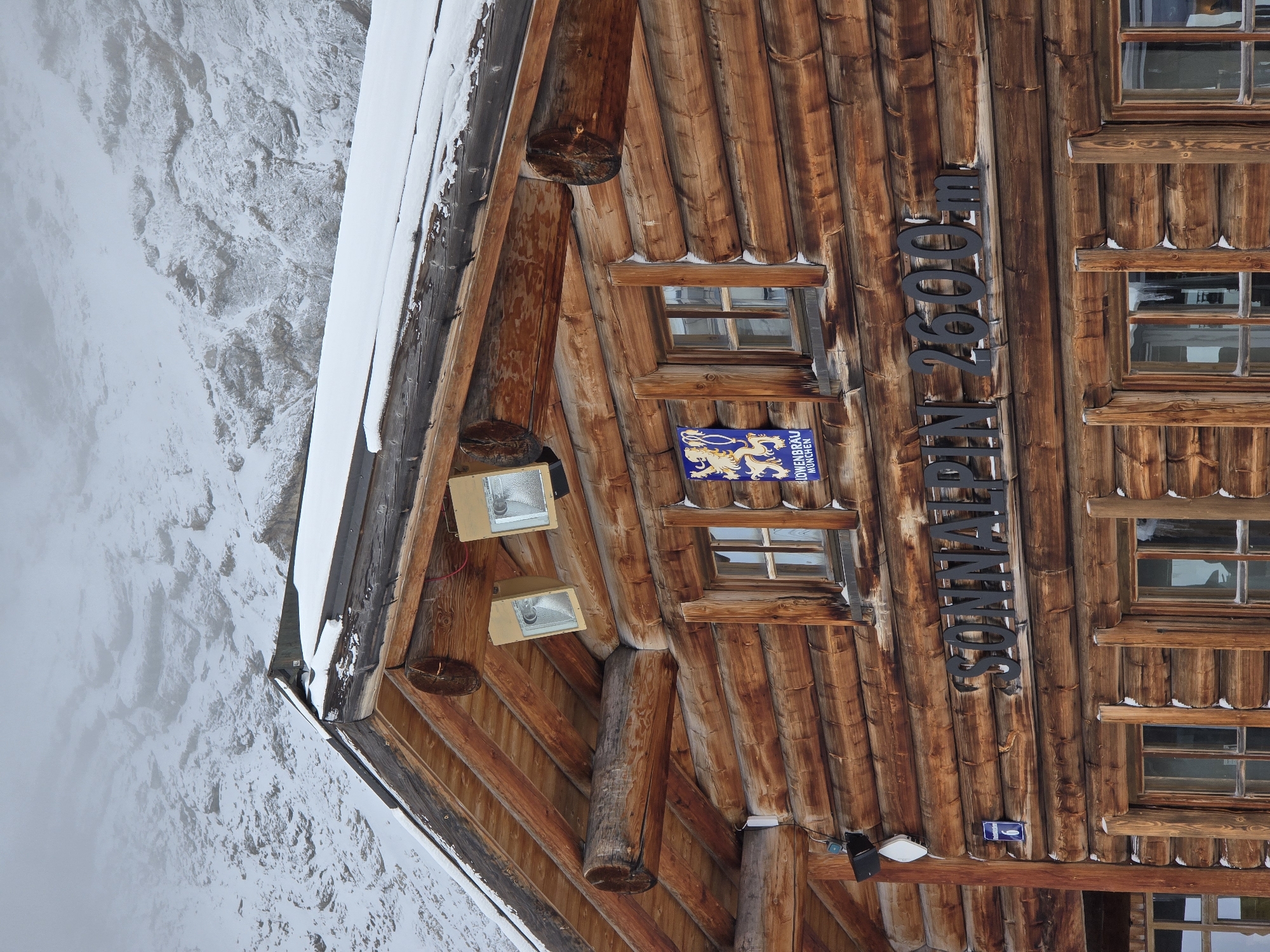 The image shows a close-up view of a rustic wooden building, likely a mountain hut, in Garmisch-Partenkirchen, Germany. The building is constructed from large, rough-hewn logs, with snow visible on the roof and in patches on the walls. Two outdoor lights are mounted high on the wooden facade. A small blue and gold emblem featuring a lion and the text "LÖWENBRÄU MÜNCHEN" is affixed to the wall near a window. Below this emblem, large black metal letters spell out "SONNALPIN 2600m". Several windows with wooden frames are visible, some with icicles hanging from the sills. The background is obscured by heavy fog or clouds, with a snowy, mountainous landscape faintly discernible through the mist. No people are visible in the image. The overall scene suggests a cold, wintery mountain environment.