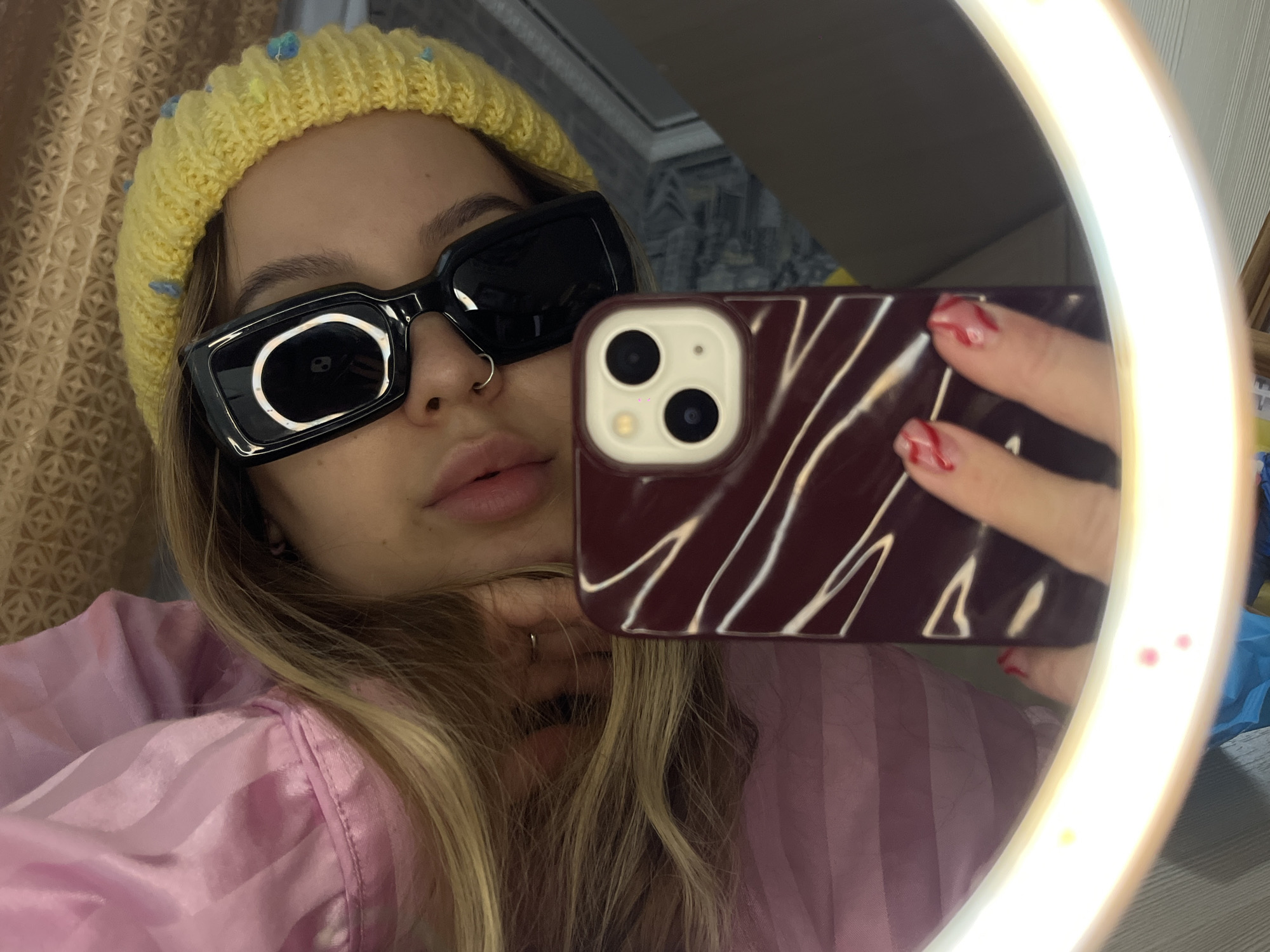 A young woman is taking a selfie in a room. She is wearing a yellow knit hat with blue accents, oversized black sunglasses, and a light pink, striped shirt. She has blonde hair and a nose ring. She is holding a dark red smartphone with white lines on the case, obscuring part of her face. The phone's camera lenses are visible. A ring light surrounds the phone. The location in the city of Novopokrovskaya, Russia is not directly determined. The woman's nails are painted with a red pattern.