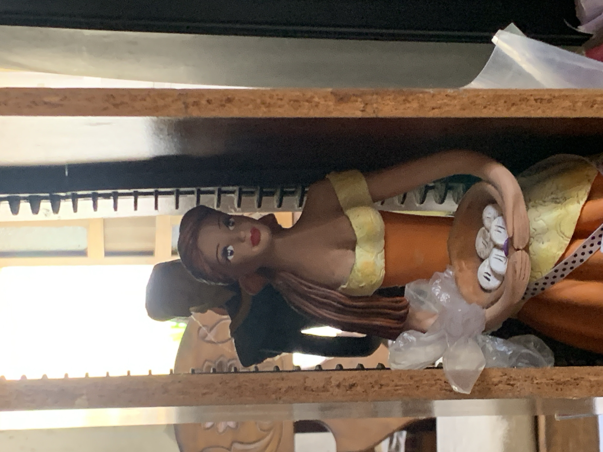 The image shows a close-up of a figurine of a woman with brown hair, wearing a yellow and orange dress. She is holding a basket containing several small, white, round objects with faces drawn on them. To her left, a piece of crinkled plastic wrap is visible. Behind her, the silhouette of another figure is discernible. The figurine is positioned within a wooden frame, which appears to be part of a shelf or display unit. The background is softly lit, suggesting an indoor setting with natural light entering from a window or doorway. The scene is devoid of any specific context that would indicate its location in Santa Ana, Venezuela.