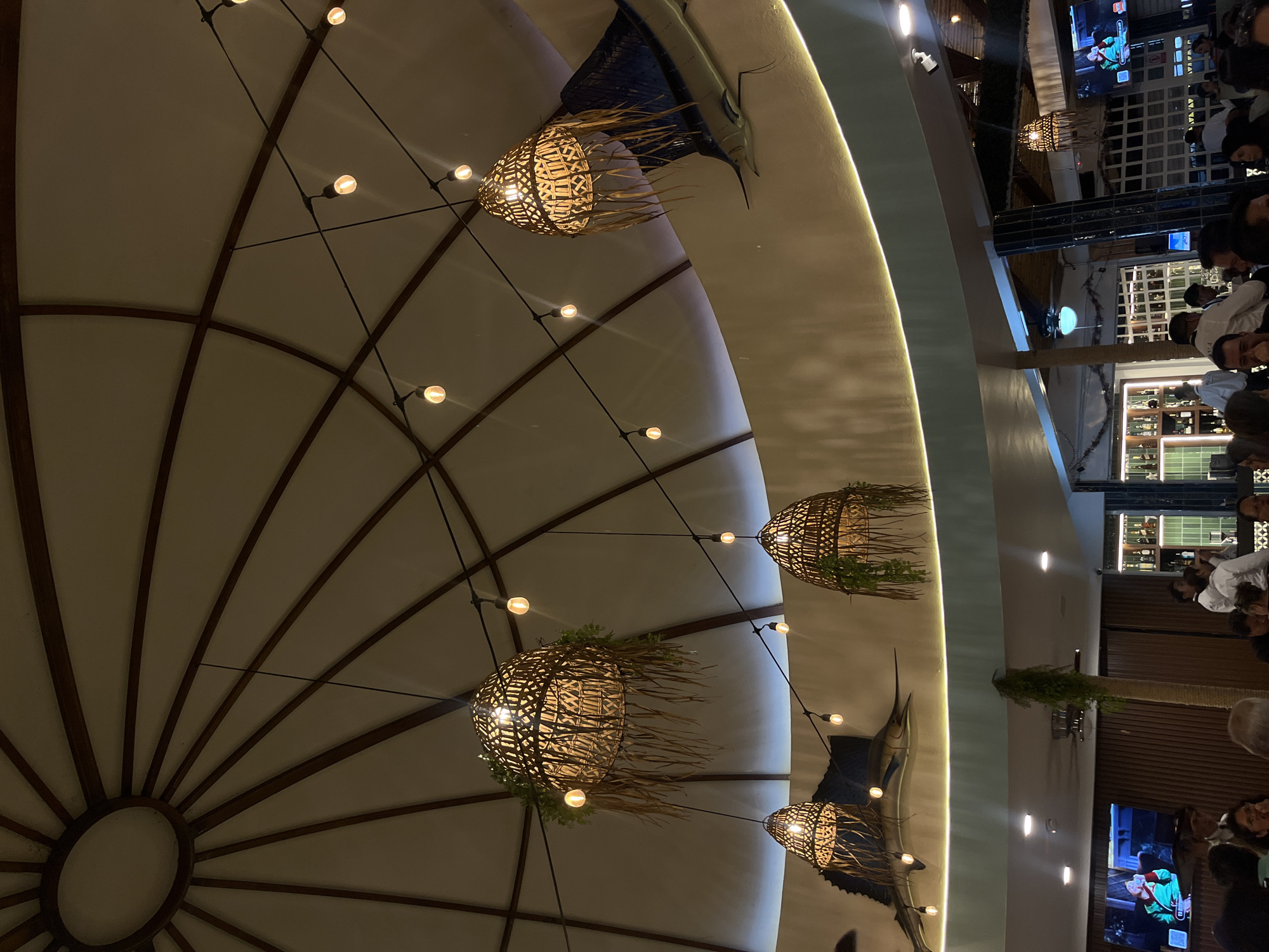 The image captures an interior scene, possibly a restaurant, located in San Bernardino Tlaxcalancingo, Mexico. The primary focal point is the elaborate ceiling design, featuring a radial pattern of dark wooden beams against a light-colored surface, with decorative straw-like lamps. String lights are strung across the ceiling. Additionally, there are taxidermied sailfish mounted on the walls. The restaurant appears to be bustling with people dining and interacting, including bartenders behind the bar. There are television sets.