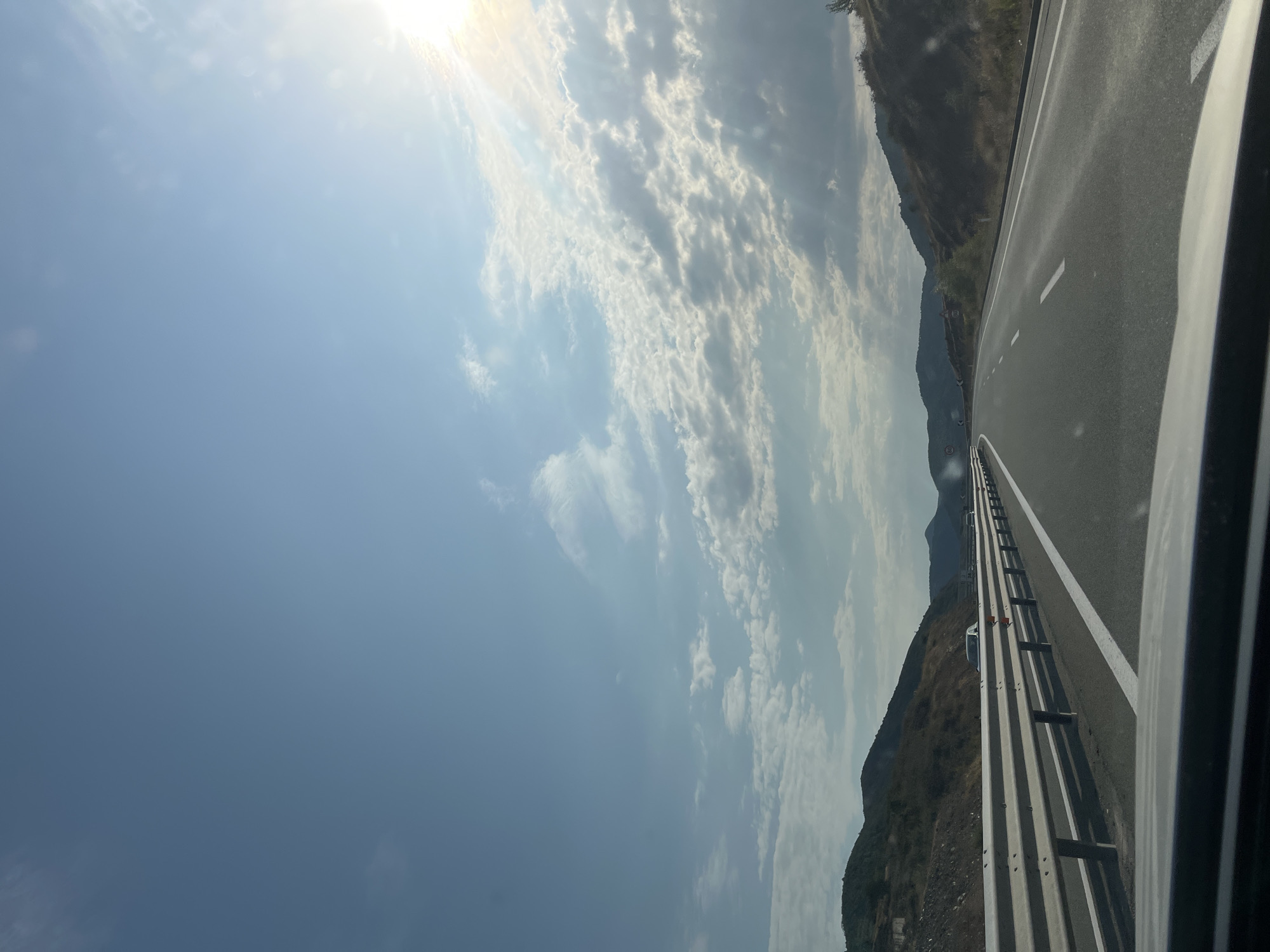 A wide-angle, eye-level shot captures a highway scene with a clear blue sky dotted with white, scattered clouds. The sun is visible on the right side of the frame, casting rays through the clouds. The highway stretches into the distance, flanked by a metal guardrail on the left and rolling hills covered in vegetation on both sides. Two cars are visible on the highway in the middle distance. The image appears to be taken from inside a vehicle, with the hood of the car visible in the foreground. The scene is consistent with a highway in a rural or semi-rural area, potentially near Rrëshen, Albania, given the landscape and road infrastructure.