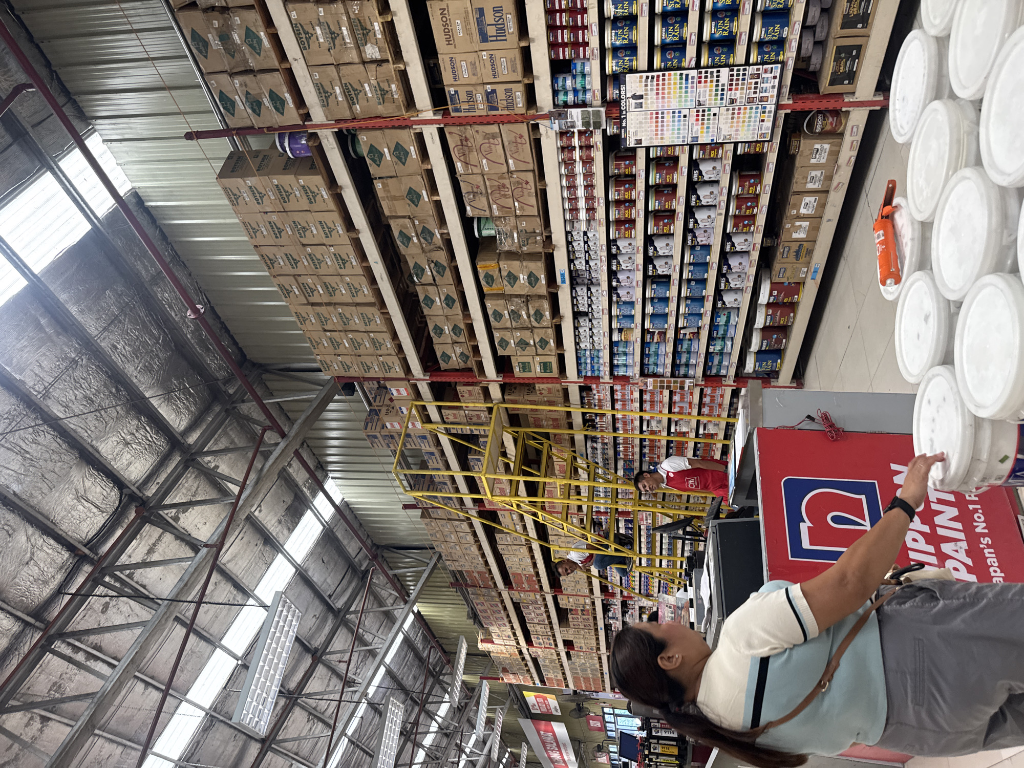 The image depicts the interior of a large hardware store, specifically a section dedicated to paint supplies. The store features tall, industrial-style shelving units stocked with various brands of paint cans, buckets, and boxes. The ceiling is high and constructed with exposed metal beams and corrugated metal, characteristic of warehouse-style buildings. Fluorescent lighting fixtures are suspended from the ceiling, supplementing the natural light coming through translucent panels.
In the foreground, a woman is partially visible from the back, wearing a light blue and white striped shirt and grey pants. She is standing near a counter with a prominent red display that has the "Nippon Paint" logo. Another man, wearing a red apron with a "CH" logo and a white shirt, is standing behind a counter to the right. A yellow ladder with multiple steps is positioned in the middle aisle, with a man in a white shirt and blue jeans sitting on one of the rungs.
The shelves are filled with rows of paint products. Some notable brands visible include "Boysen," "Hudson," "Davies," and "Nippon Paint." There is also a color swatch display. In the lower right quadrant of the image, several white buckets are neatly arranged, some of which appear to contain white paint. An orange paint roller and extension pole are resting on top of some of these buckets. The store appears well-organized and fully stocked.
The sign on the left side of the image reads "DAKANG SALAMAT," a Filipino phrase meaning "Thank you very much." Based on the presence of "Nippon Paint," a prominent brand in the Philippines, and the overall aesthetic of the store, it is likely located in the Philippines. This specific store is the Ace Hardware located in the Gaisano Mall of Tagum, Philippines. The store's interior aligns with the typical layout and branding of Ace Hardware stores in the Philippines, which often include a dedicated paint section with prominent displays of brands like Nippon Paint. The combination of the store's architecture, product offerings, and signage strongly suggests this location.