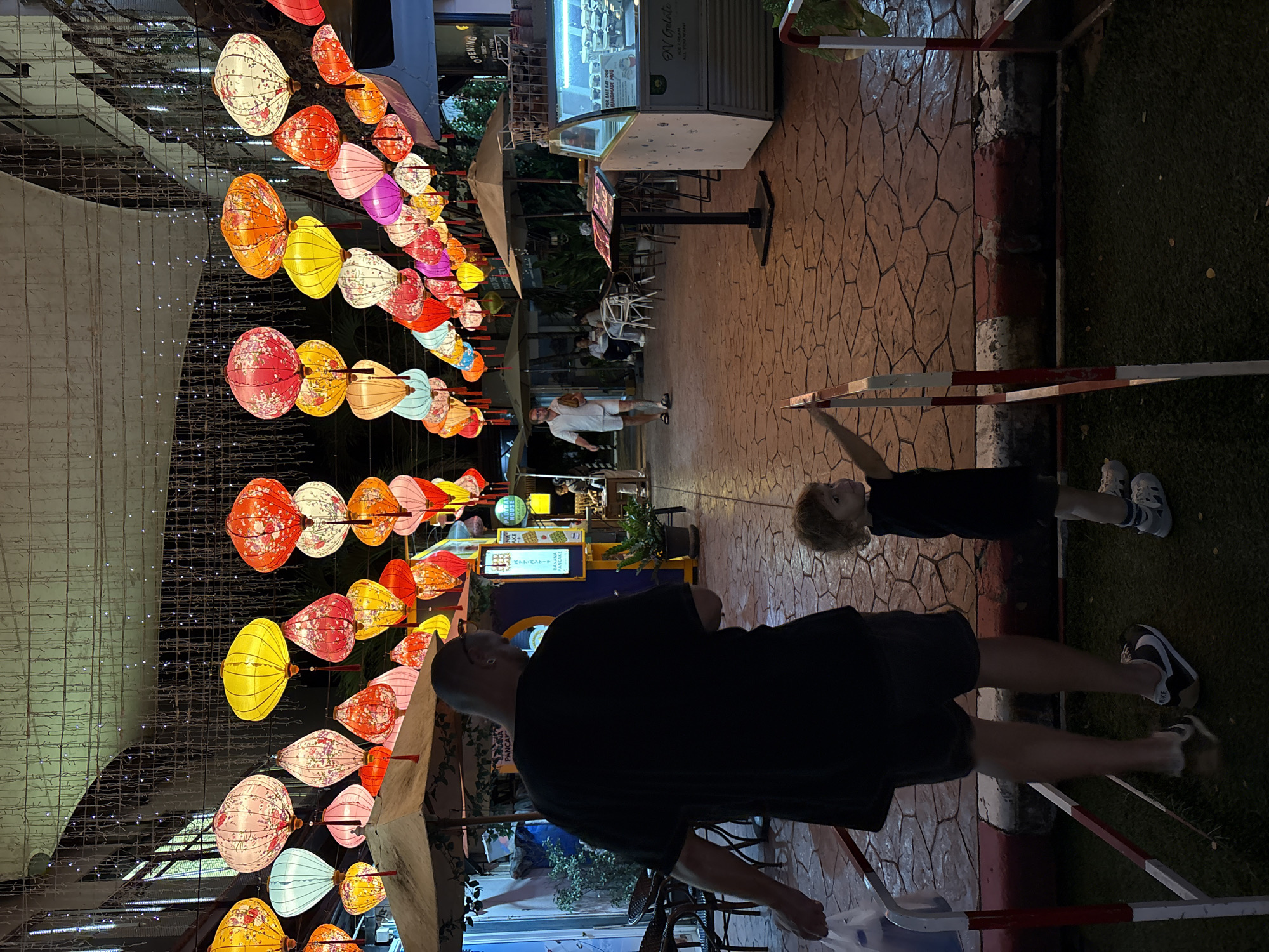 In Choeng Thale, Thailand, an evening scene unfolds under a canopy of vibrant, illuminated paper lanterns strung across a pedestrian walkway. A man with a shaved head, wearing a black t-shirt and dark shorts, walks away from the camera, holding a white plastic bag. Beside him, a young child, likely a toddler, stands facing away from the viewer, reaching up towards the lanterns. The child wears a dark t-shirt and shorts, with white sneakers and socks. The walkway is paved with hexagonal stones, and to the right, an ice cream display case is visible. In the background, several people are seated at tables beneath umbrellas, and shops line the street. The atmosphere is festive and illuminated by the soft glow of the lanterns and surrounding lights.