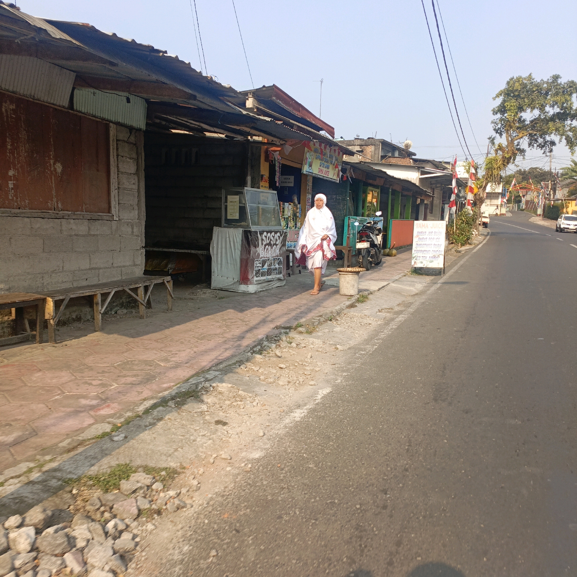 A street-level view of a row of shops on the side of a paved road in Salatiga, Indonesia. The shops appear to be small, open-fronted structures with corrugated metal roofs. One shop has a display case filled with goods, and a sign that reads "SOSIS BAKAR" (grilled sausage) and "JUICE BUAH SEGAR" (fresh fruit juice). A person dressed in white, possibly a religious garment, is walking on the sidewalk in front of the shops. Another shop has a sign advertising "TAMA JUICE" with a list of drinks. A motorcycle is parked in front of one of the shops. Flags and banners, some with red and white stripes, are visible along the street, suggesting a local celebration or event. The road is asphalt and appears to be in moderate condition. A white car is visible further down the road. The environment is sunny with clear blue skies.
