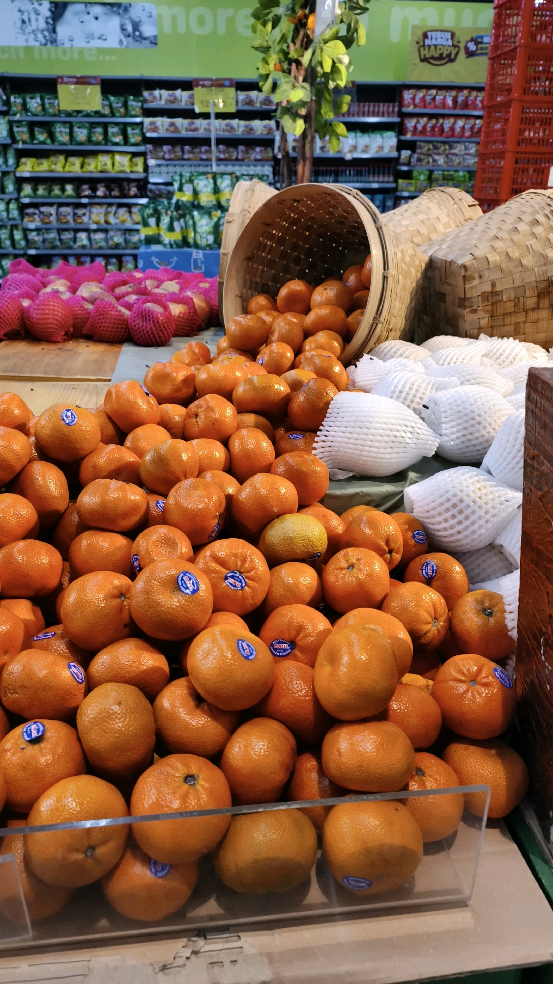 The image depicts a produce display at a supermarket, likely in Baturaja, Indonesia, given the text visible on a sign that says "Happy" and the general aesthetic of a Indonesian grocery store. In the foreground, a large pile of fresh, bright orange tangerines is displayed, some of which have blue stickers on them. These tangerines are spilling out of a large, woven bamboo basket that is tilted on its side. To the right of the tangerines, several white foam-wrapped fruits, possibly pears or apples, are neatly arranged. In the background, shelves are stocked with various packaged goods, and a decorative tree with green leaves stands behind the fruit display. Red plastic crates are visible on the far right. The overall impression is of a vibrant and abundant fresh produce section.