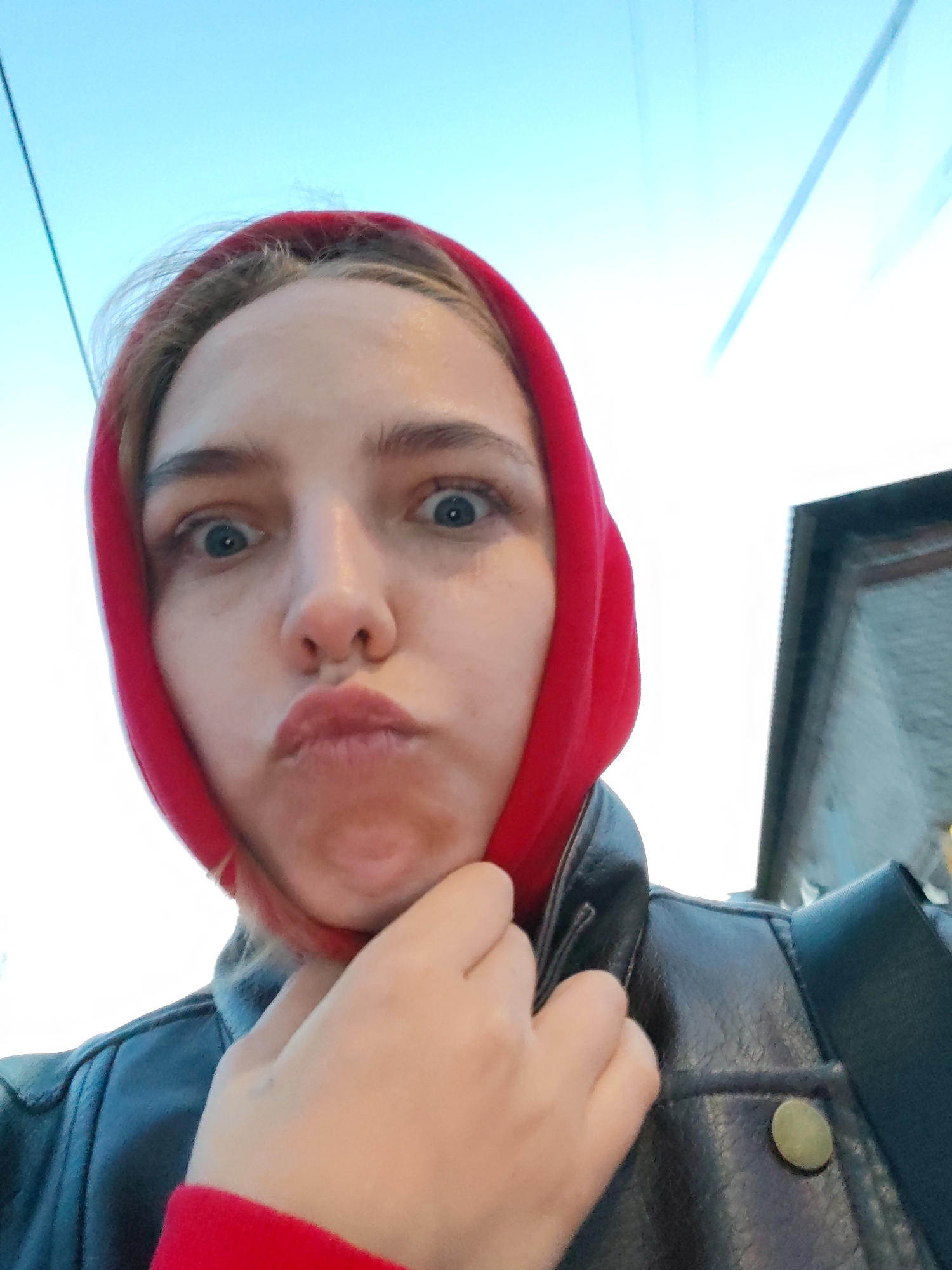 A young woman is captured in a close-up, upward-angled selfie, exhibiting a playful and exaggerated expression. Her blue eyes are wide open and directly engaging the camera, while her lips are pursed in a "fish-lips" or "duck face" pose. She wears a vibrant red hood or head covering that frames her face, with strands of light brown hair visible at her forehead. A dark brown or black leather jacket covers her, and an olive-green snap is visible on the jacket's collar to her right. One of her hands is partially visible, cupping her chin.
The background consists primarily of a clear, bright blue sky, indicating daytime and good weather. Thin, dark lines, possibly wires or cables, traverse the sky in the upper left and right areas of the frame. On the far right, a dark, rectangular element, which could be part of a building or vehicle, is partially visible. The overall mood conveyed by the subject's expression is one of lightheartedness and humor. No text is visible in the image. The setting appears to be an outdoor urban or suburban environment, potentially in Chaikovsky, Russia, as suggested by the location context.